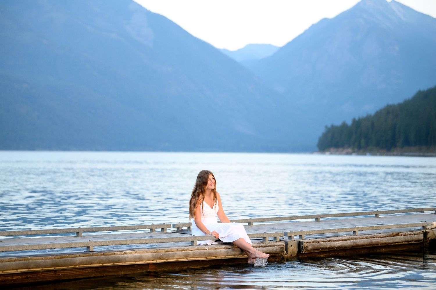 Someone in a white dress sits on a wooden dock overlooking a serene mountain lake on a misty morning.