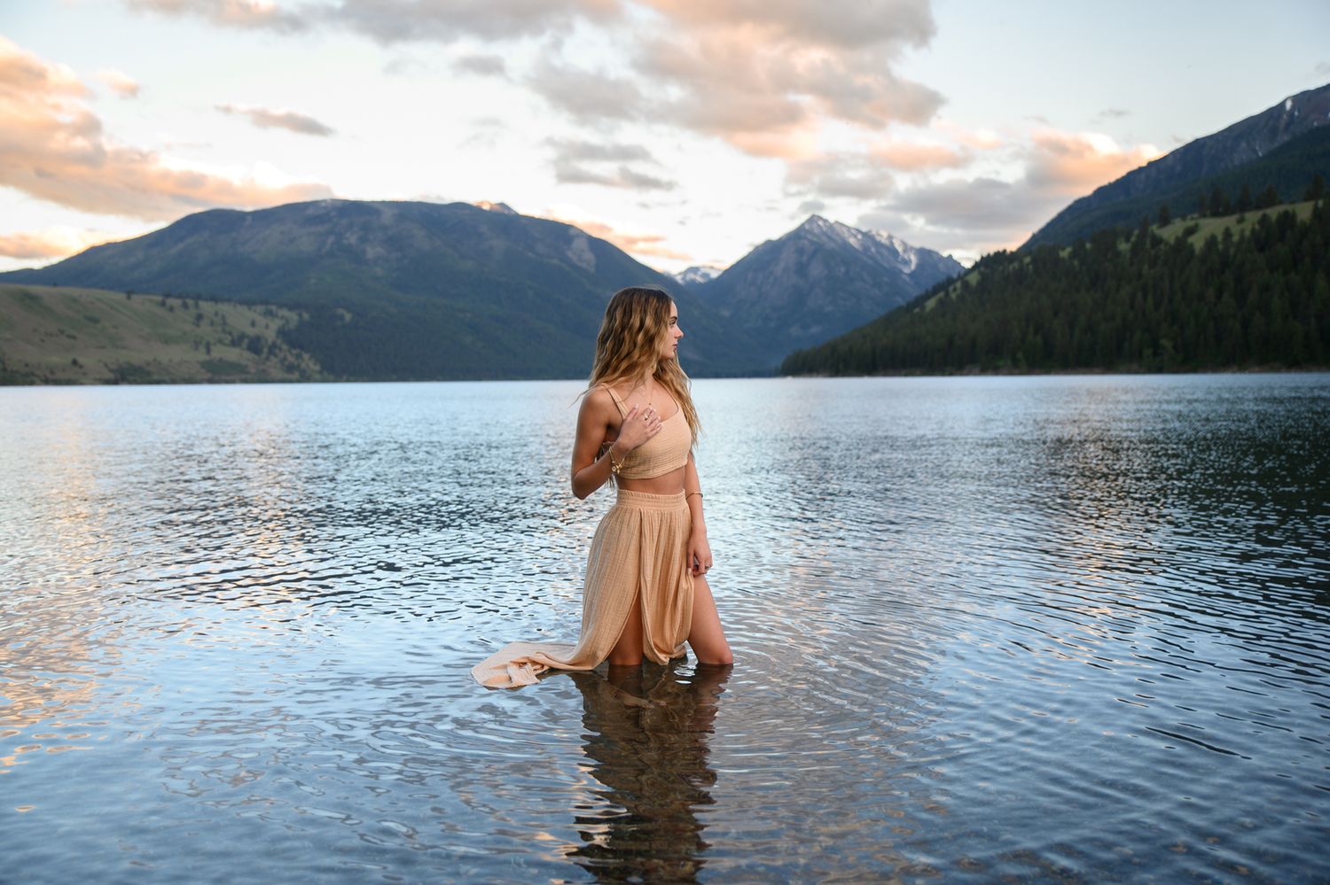 A person in a flowing peach dress stands in a mountain lake at sunset, surrounded by snow-capped peaks.