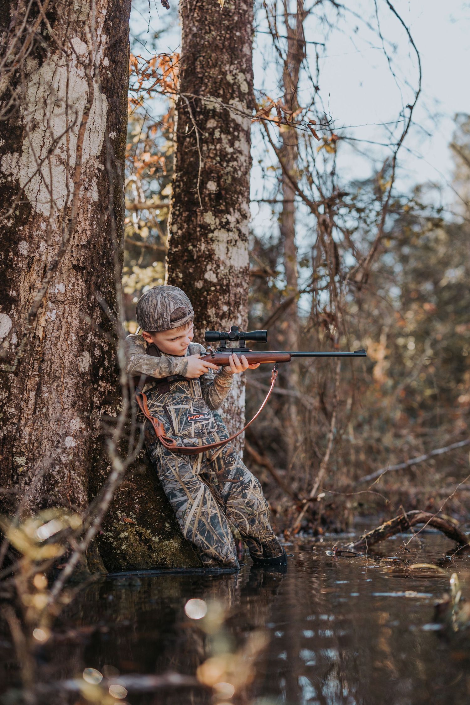 Hunter in camouflage clothing aims rifle while leaning against tree trunk in autumn forest setting.