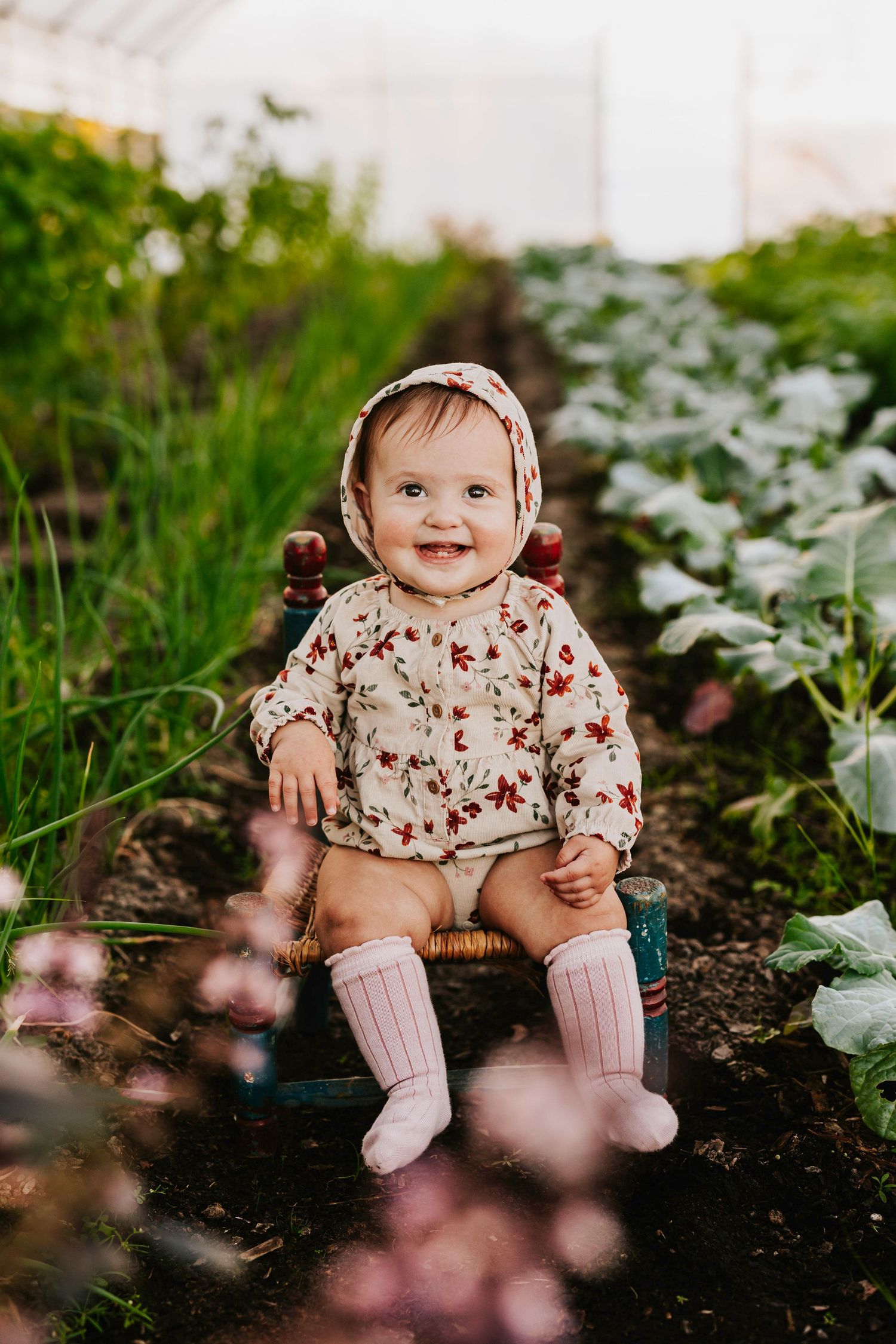 Baby in floral dress and bonnet sitting on garden stool among blooming flowers.