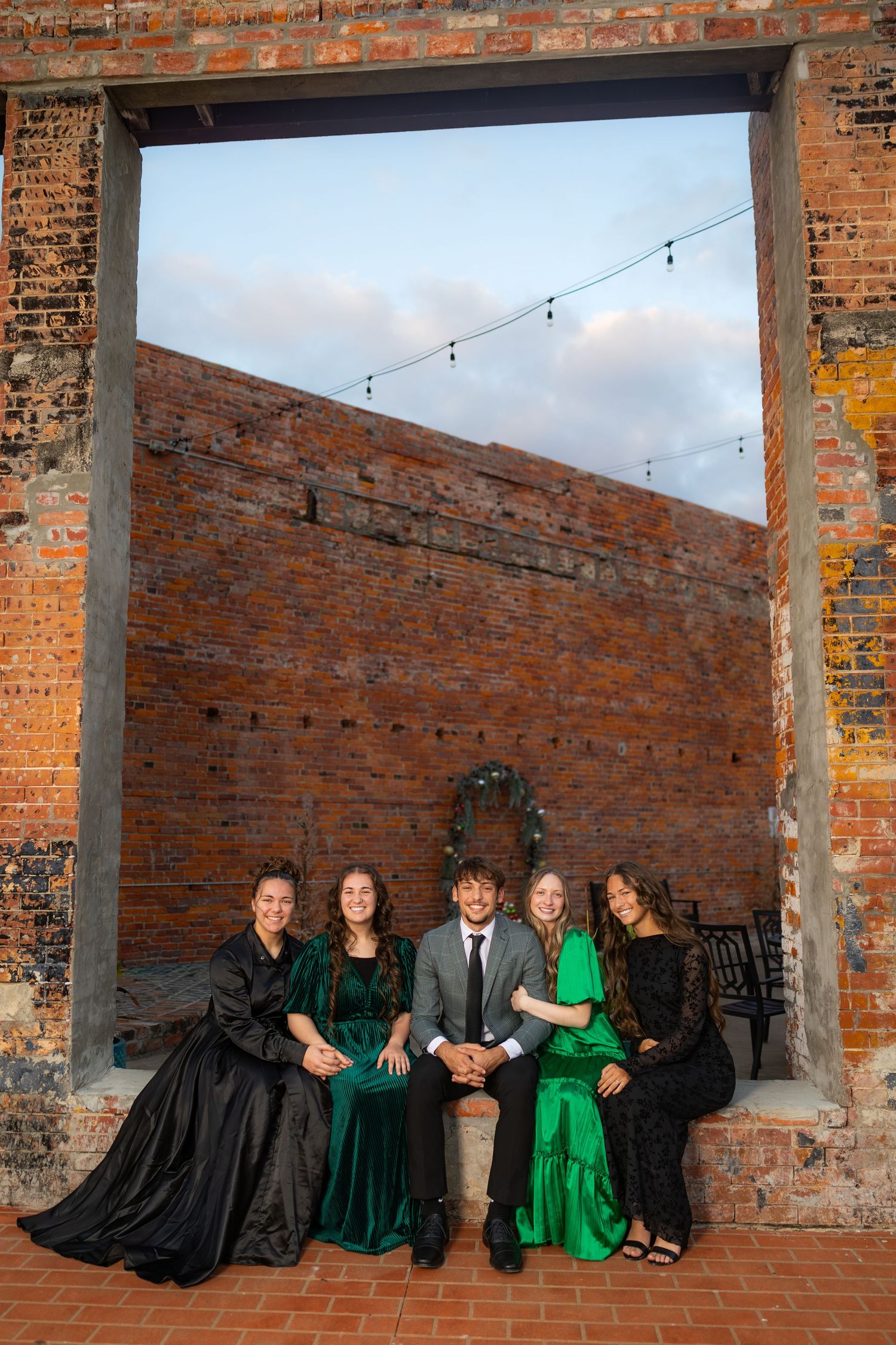 Group in formal attire lounges against brick wall with string lights overhead.