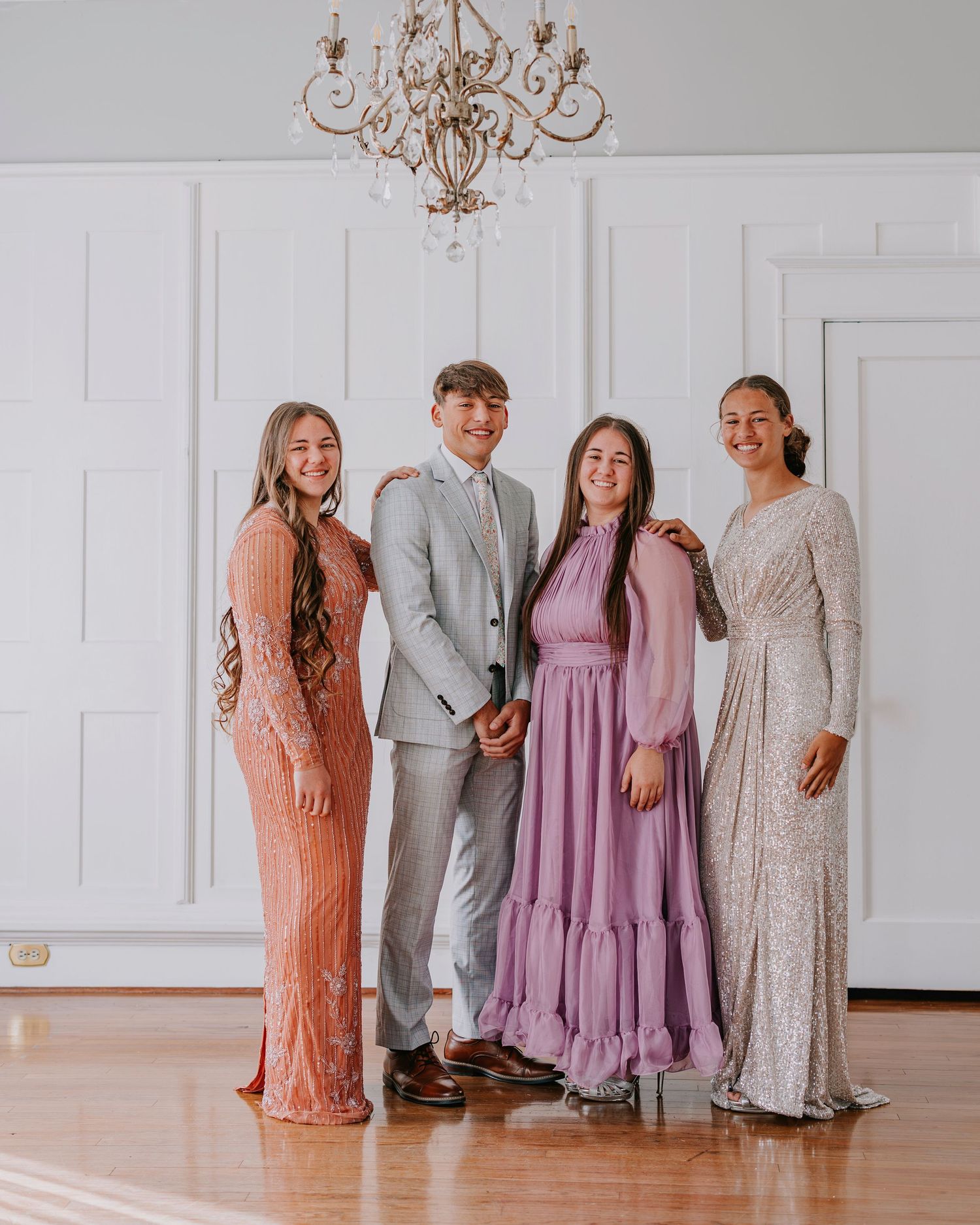 Group poses in formal attire under elegant chandelier in white room.