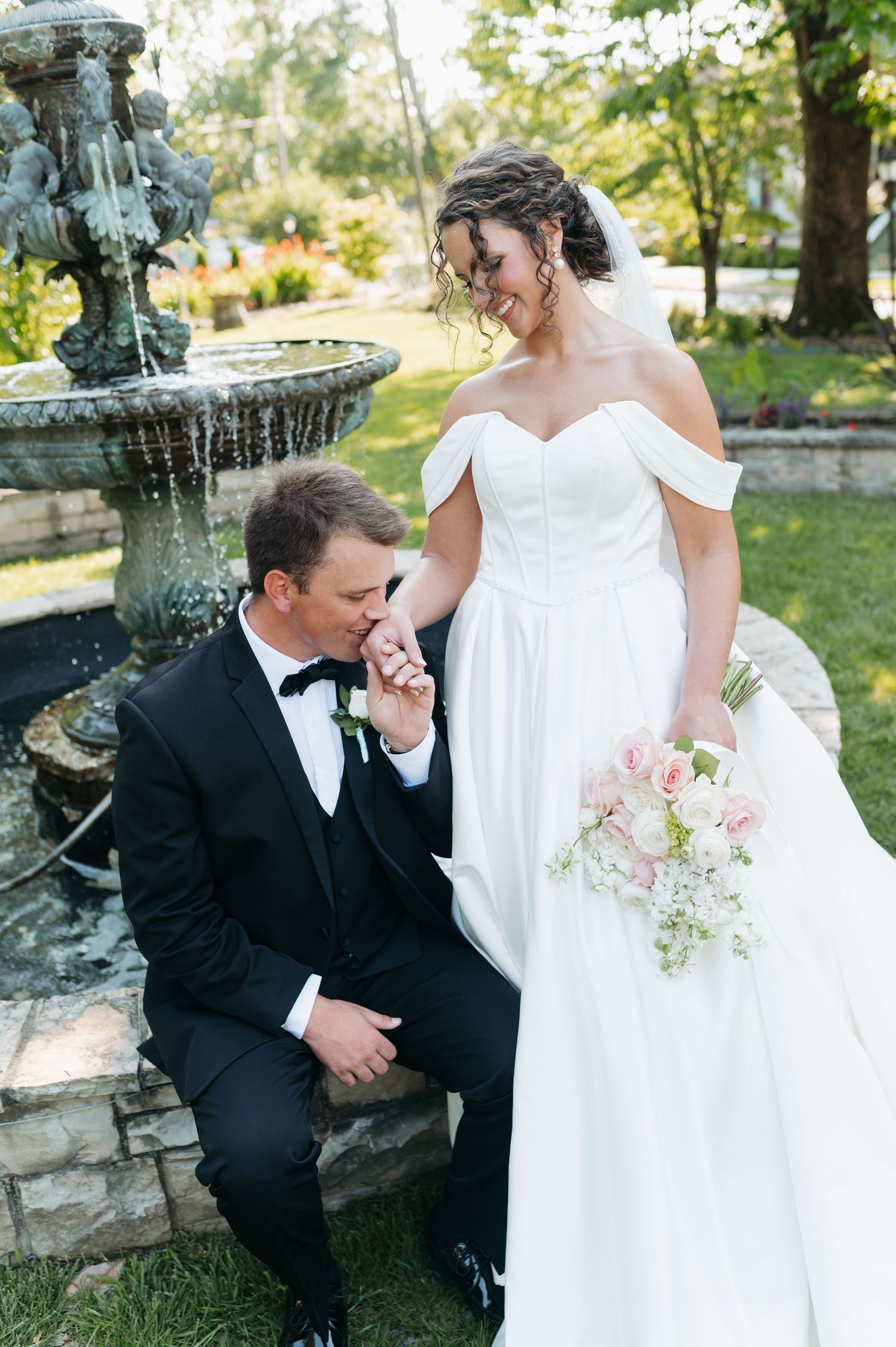 Groom in black tuxedo kisses bride's hand near stone fountain, bride holds pink bouquet in white off-shoulder gown.
