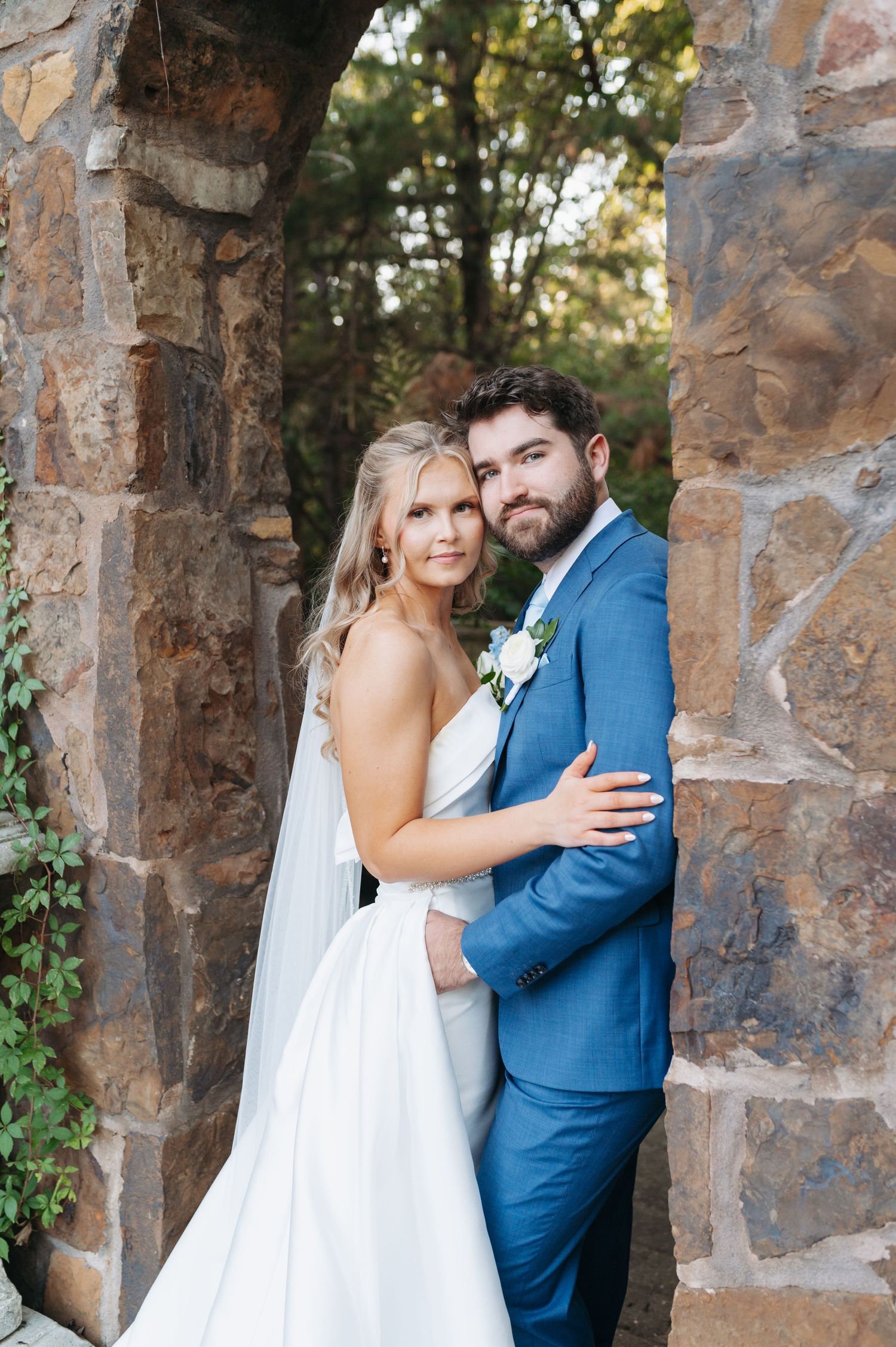 Bride and groom pose together against a stone arch, she in a white gown with veil, he in a blue suit with boutonniere.