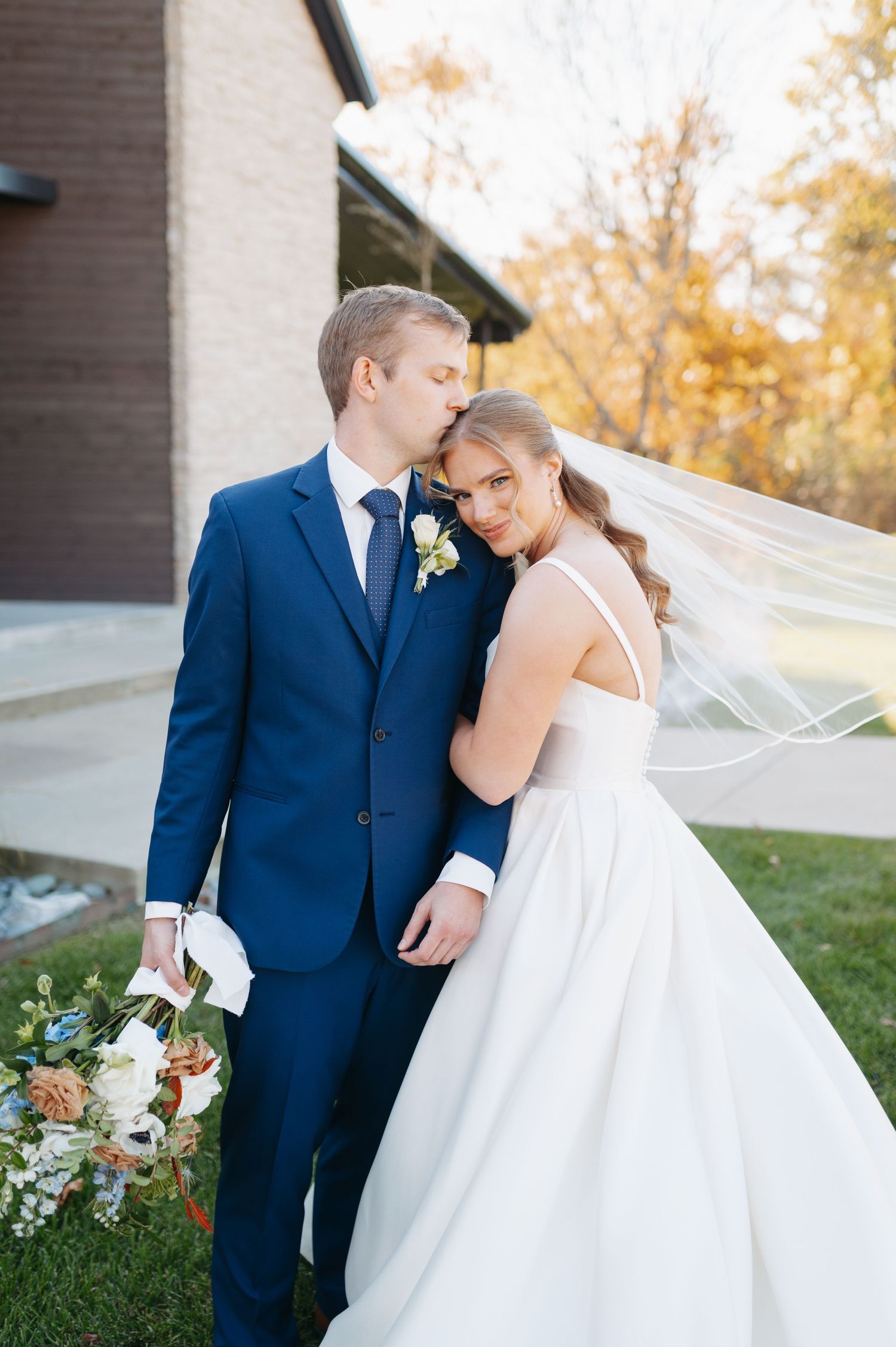 Bride in white gown with flowing veil and groom in navy suit share a tender moment outdoors at fall wedding.