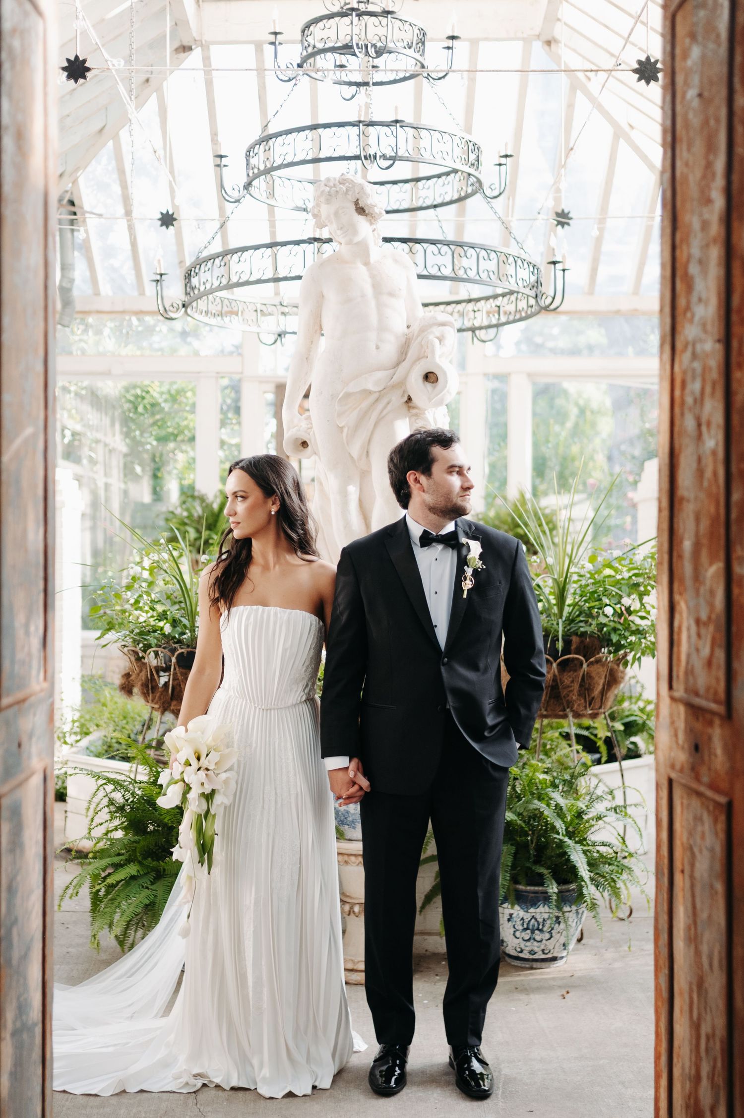 Elegant bride and groom hold hands in a glass conservatory, with a white marble statue and lush greenery behind them.