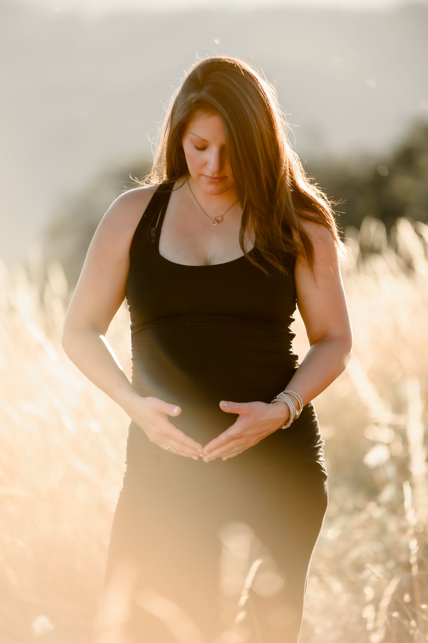 Pregnant woman in black outfit cradling her baby bump in a sunlit golden field.