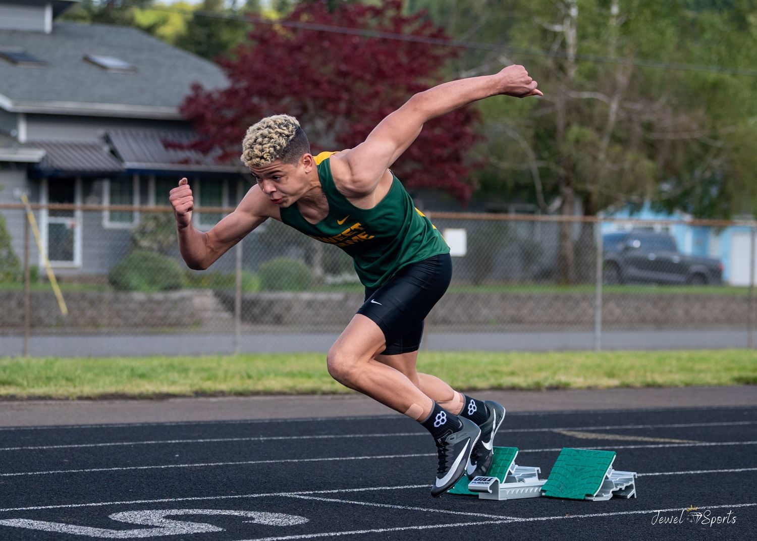 Young sprinter in green uniform exploding off starting blocks on an outdoor track.