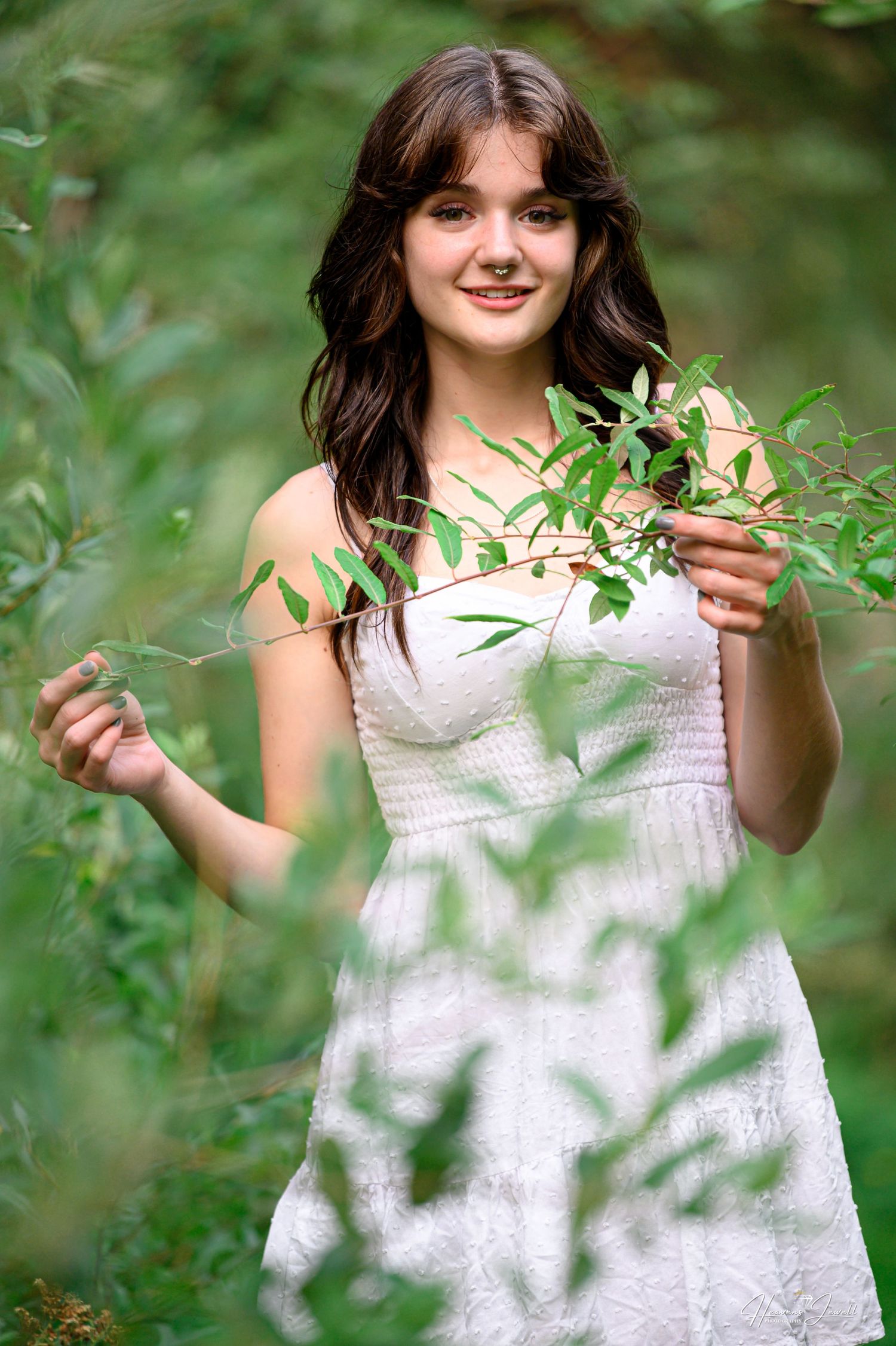 Smiling teenage girl in white sundress holding green leafy branches in lush outdoor nature setting.