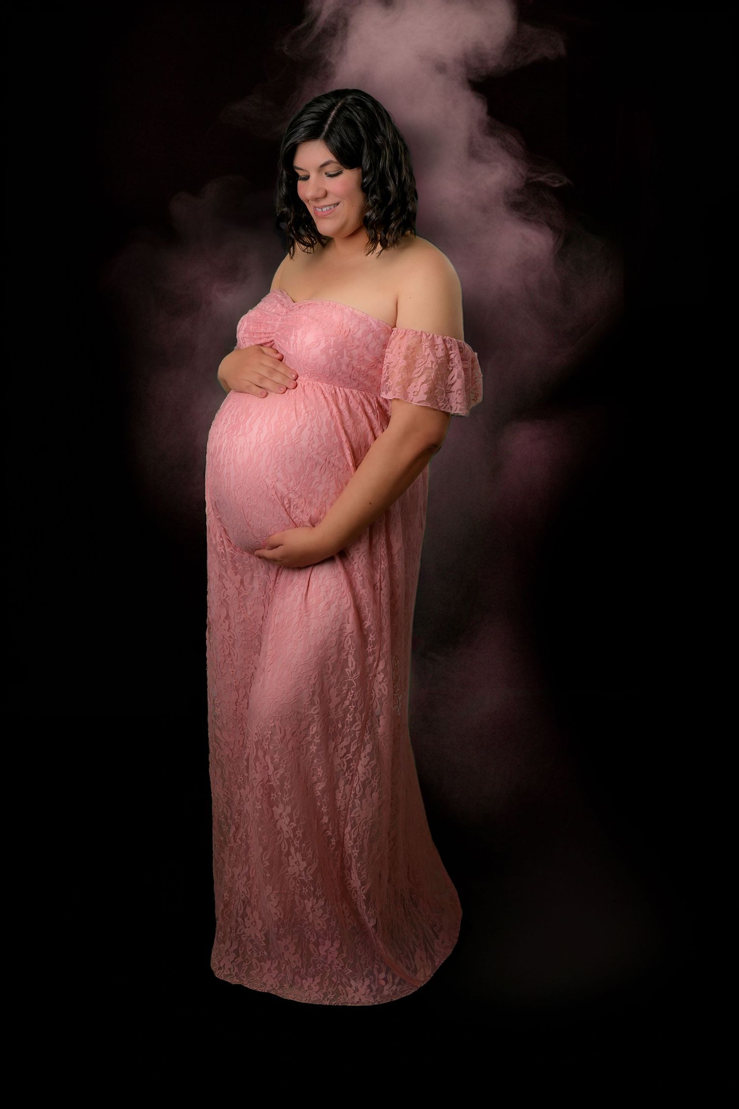 Pregnant woman in elegant pink lace off-shoulder maternity gown posing against dramatic dark smoky background.