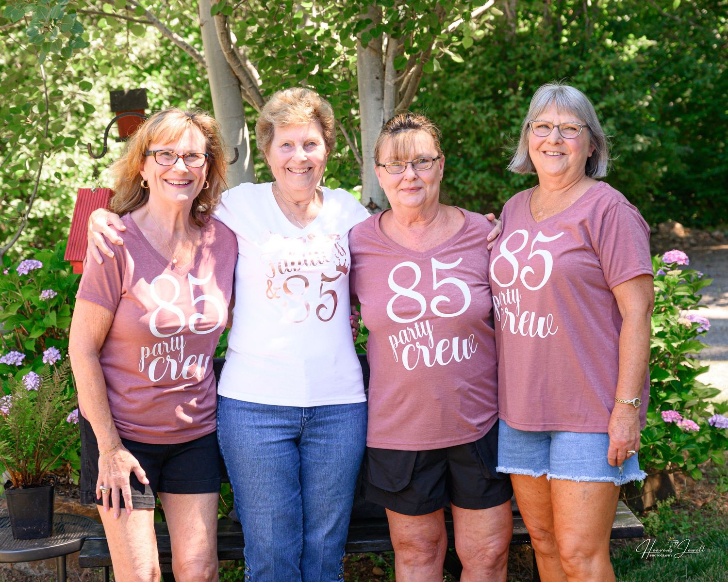 Four older women smiling outdoors wearing matching 85th birthday party crew t-shirts.