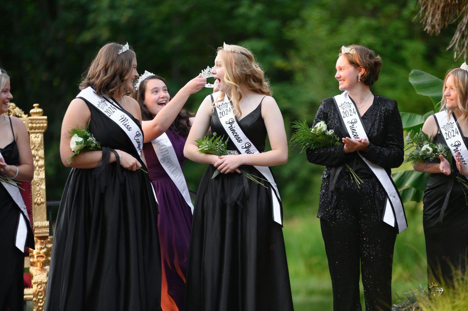 Young women in black formal gowns during outdoor pageant crowning ceremony with sashes and bouquets.
