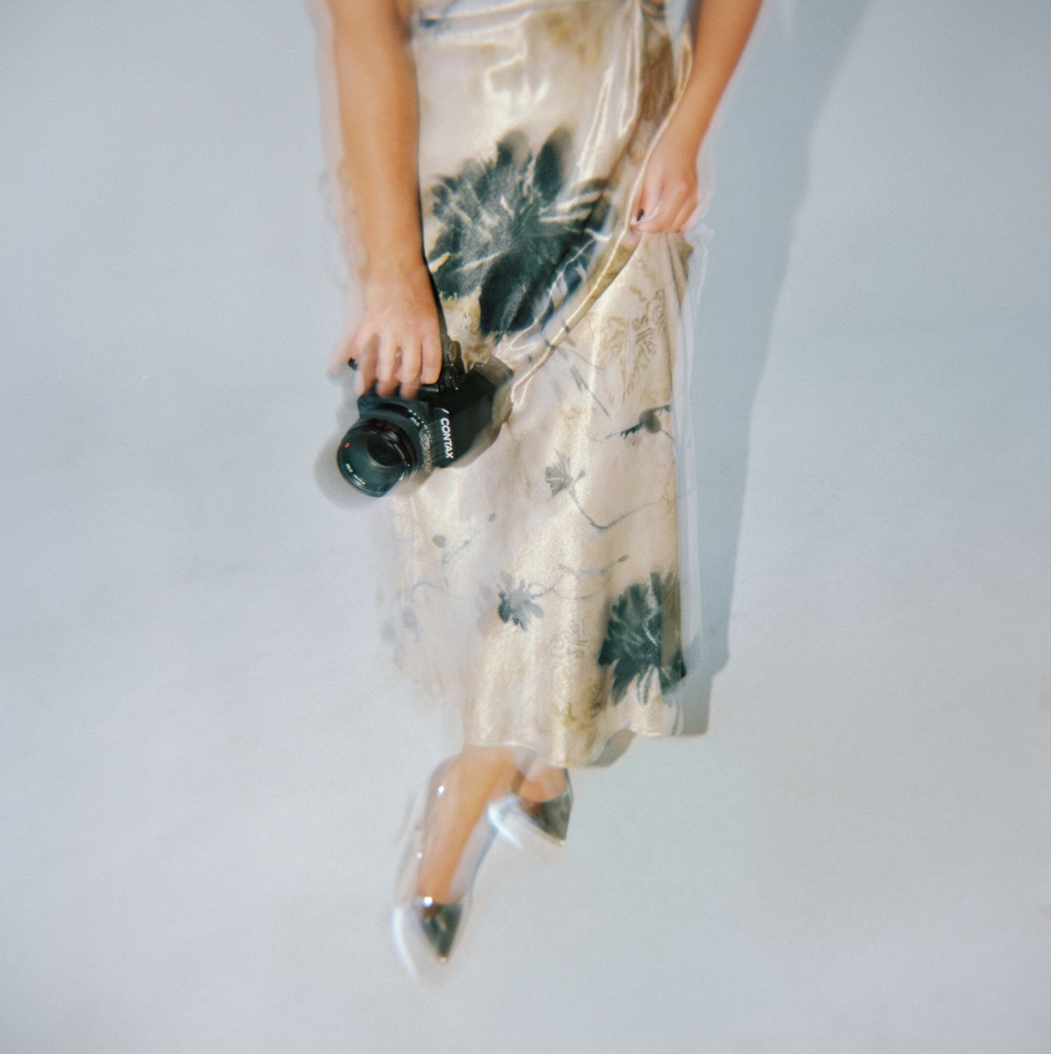 Elegant woman in cream floral dress and metallic pumps carrying a professional camera, overhead view.