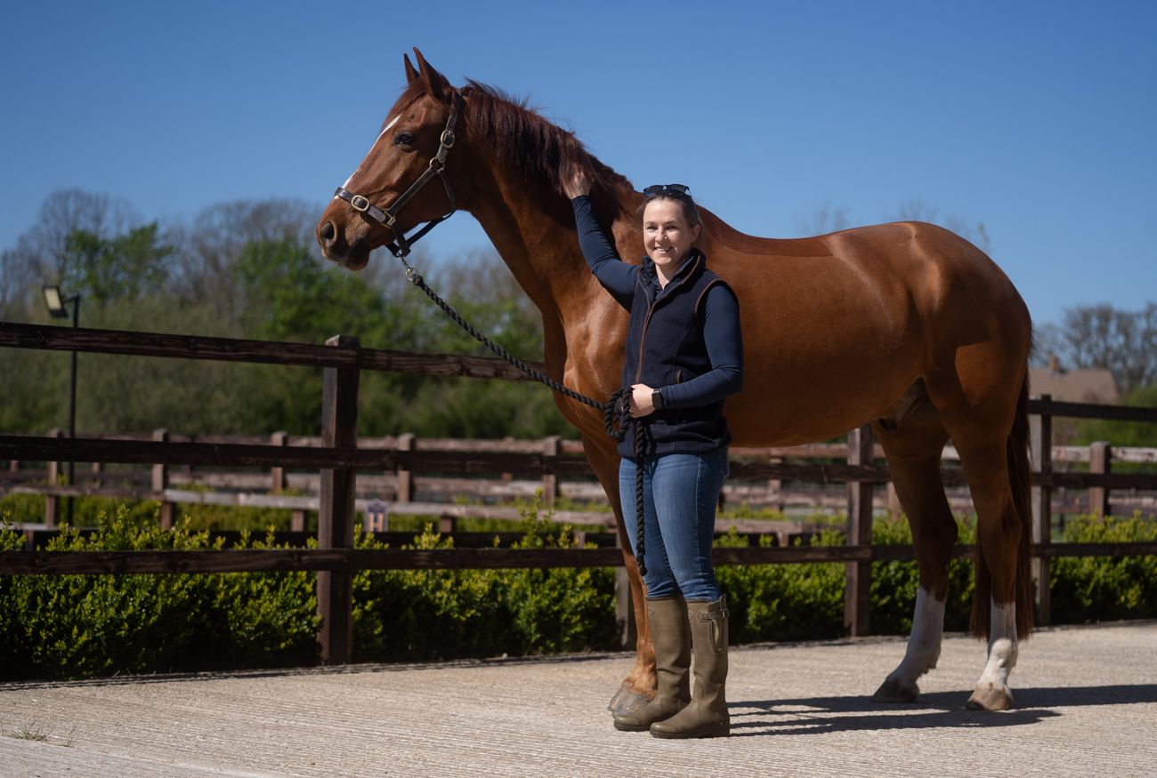 A smiling woman in riding boots stands beside a chestnut horse in a sunny equestrian arena with wooden fencing.