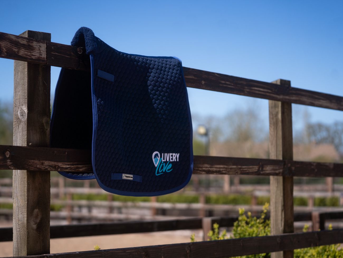 Navy blue quilted horse saddle pad with Livery Live logo displayed on a wooden fence outdoors.