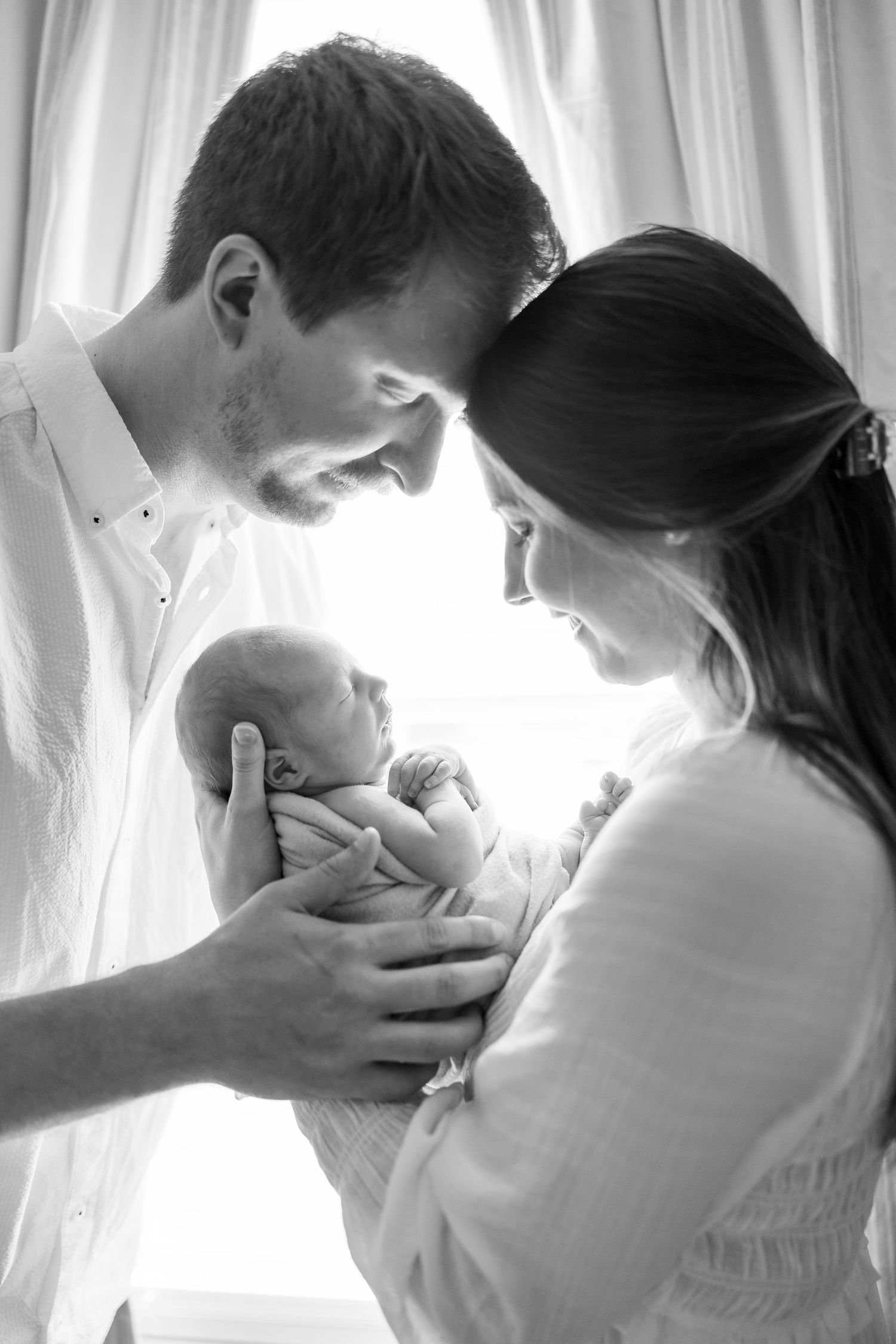 Black and white portrait of parents lovingly gazing at their newborn baby by a window.