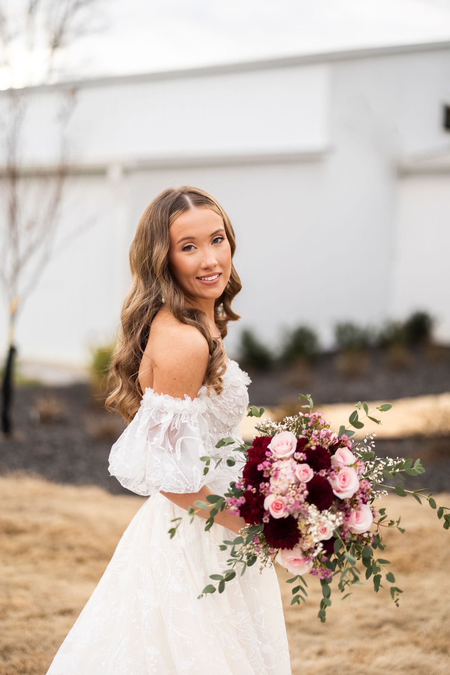 Bride in off-shoulder dress holding vibrant pink and burgundy bouquet smiles at camera.