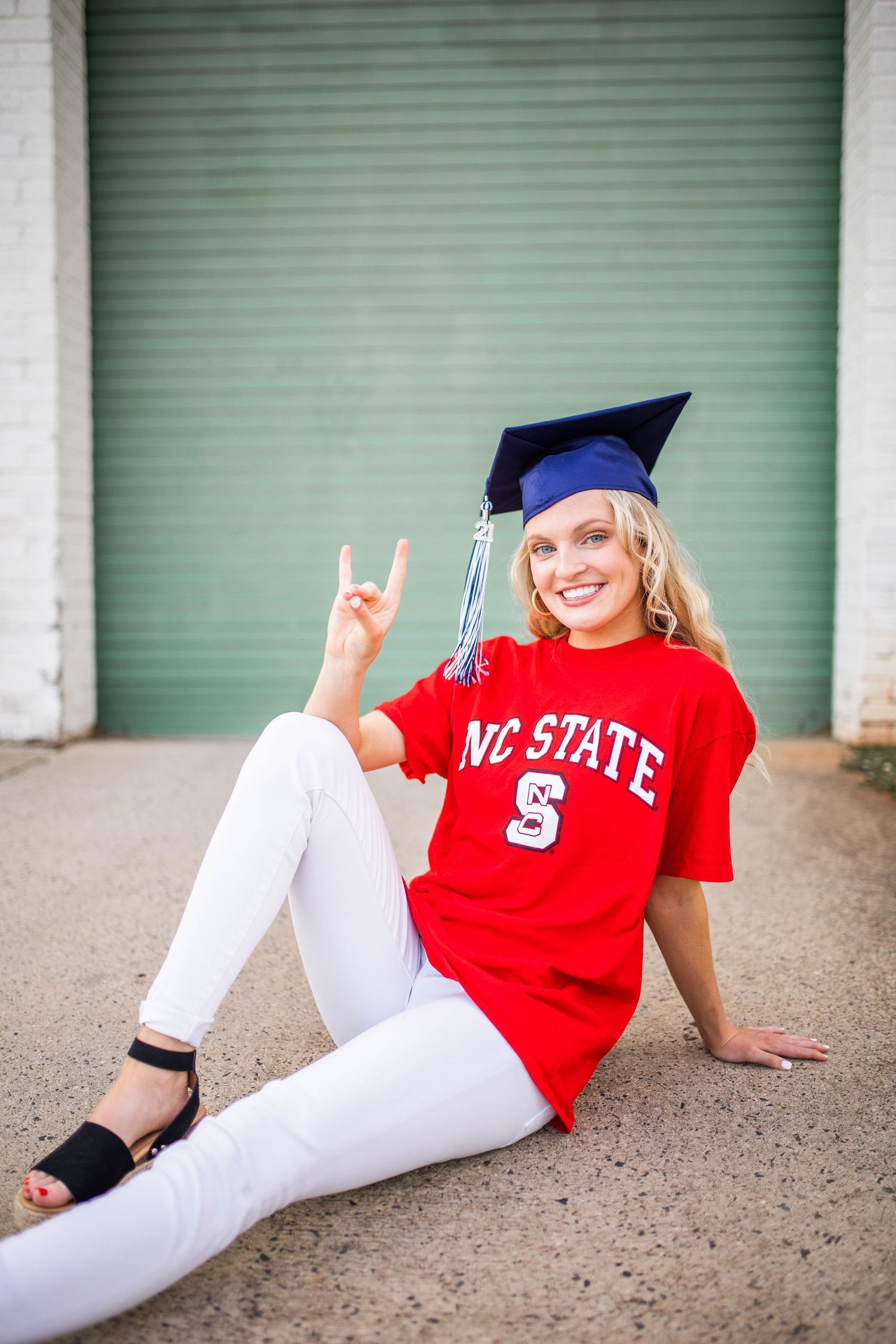 Graduate in NC State shirt and cap poses casually against green garage door.