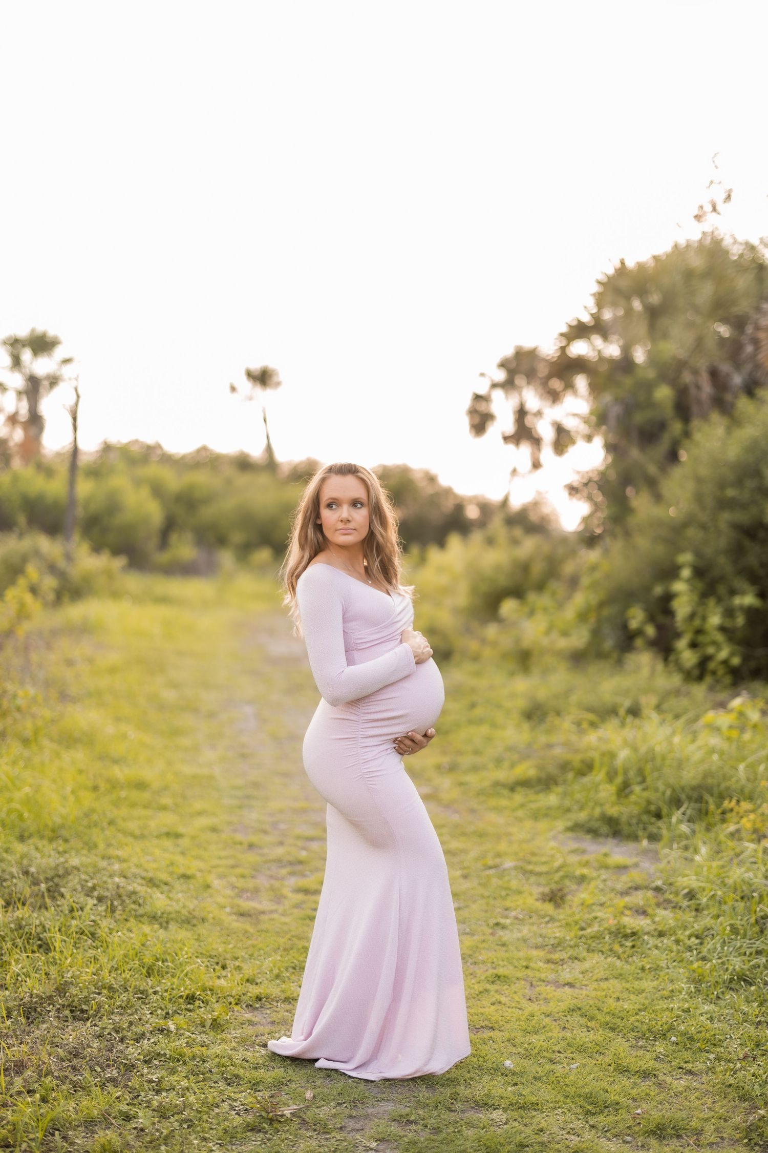 Expecting mother in a flowing pink dress poses on a nature path at sunset.