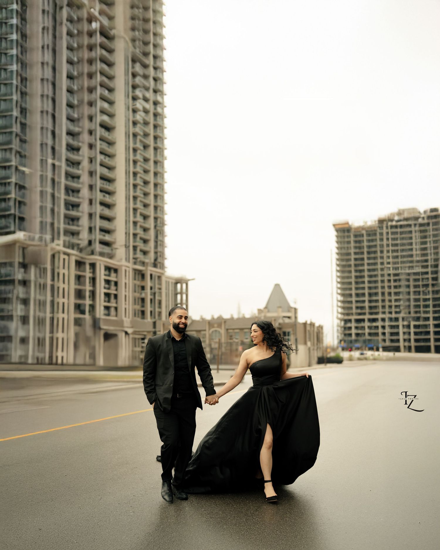 Beautiful Indian couple running hand-in-hand, dressed in elegant black outfits, looking at each other amidst urban city buildings during out