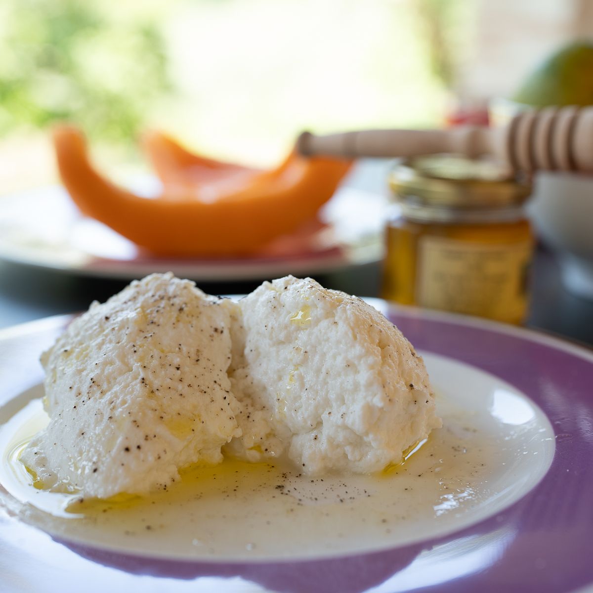 Fresh ricotta cheese drizzled with golden honey on a white plate, with a jar of honey and melon in the background.