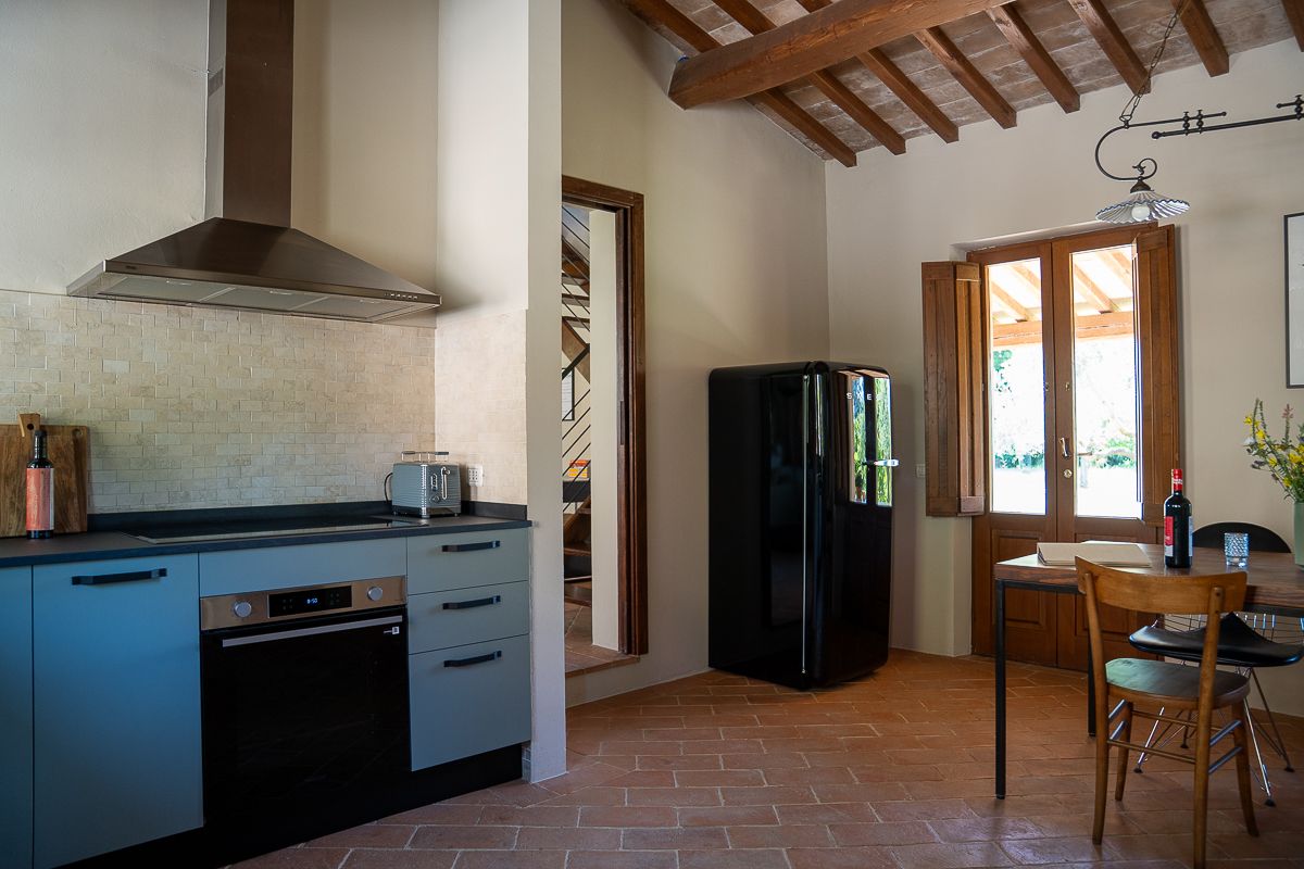 Rustic Tuscan kitchen with blue cabinets, black refrigerator, terracotta floors, wooden ceiling beams, and a dining area by a window.