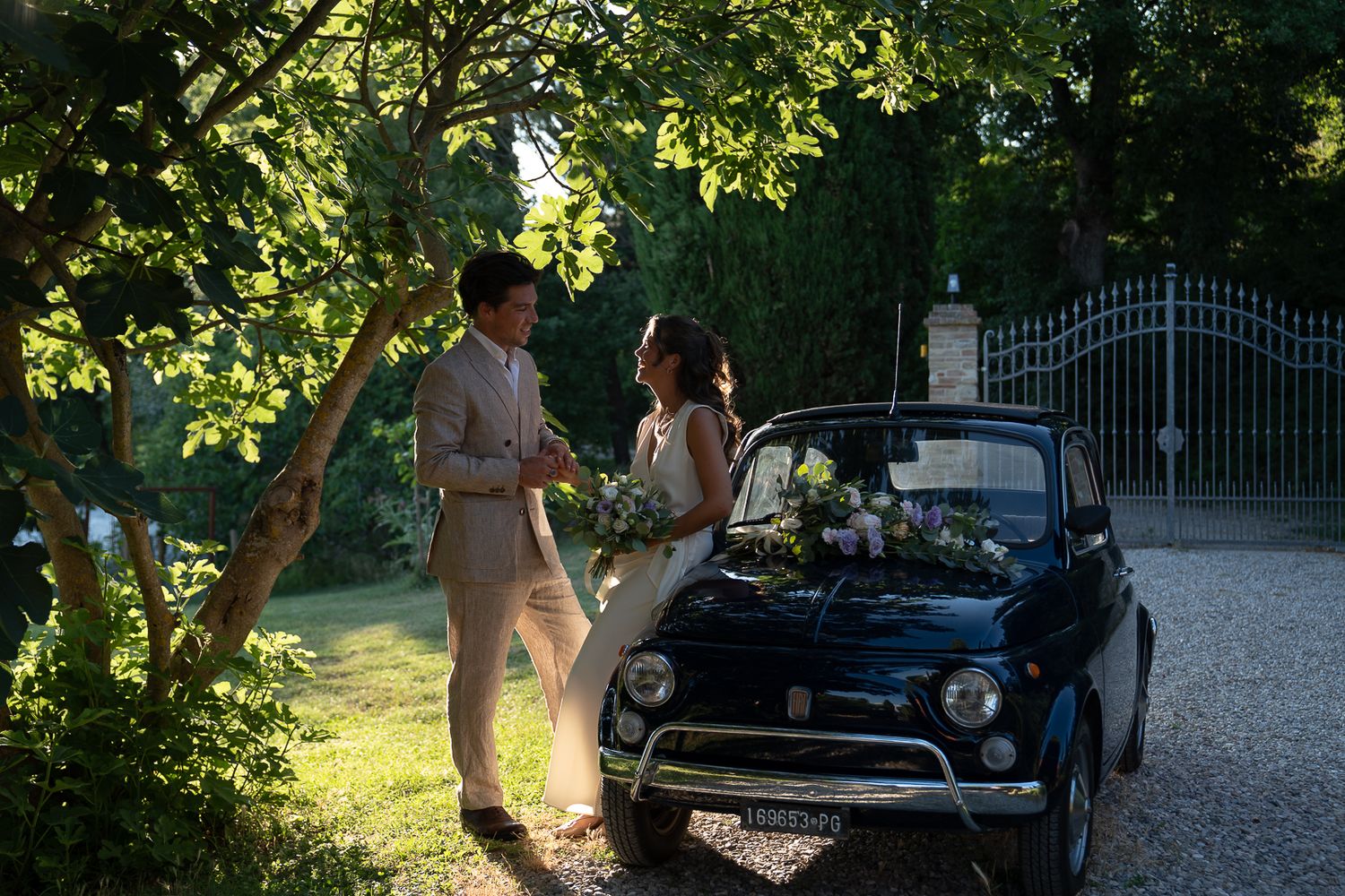 Bride and groom standing beside a vintage black Fiat 500 in a sunlit garden, holding a bouquet.
