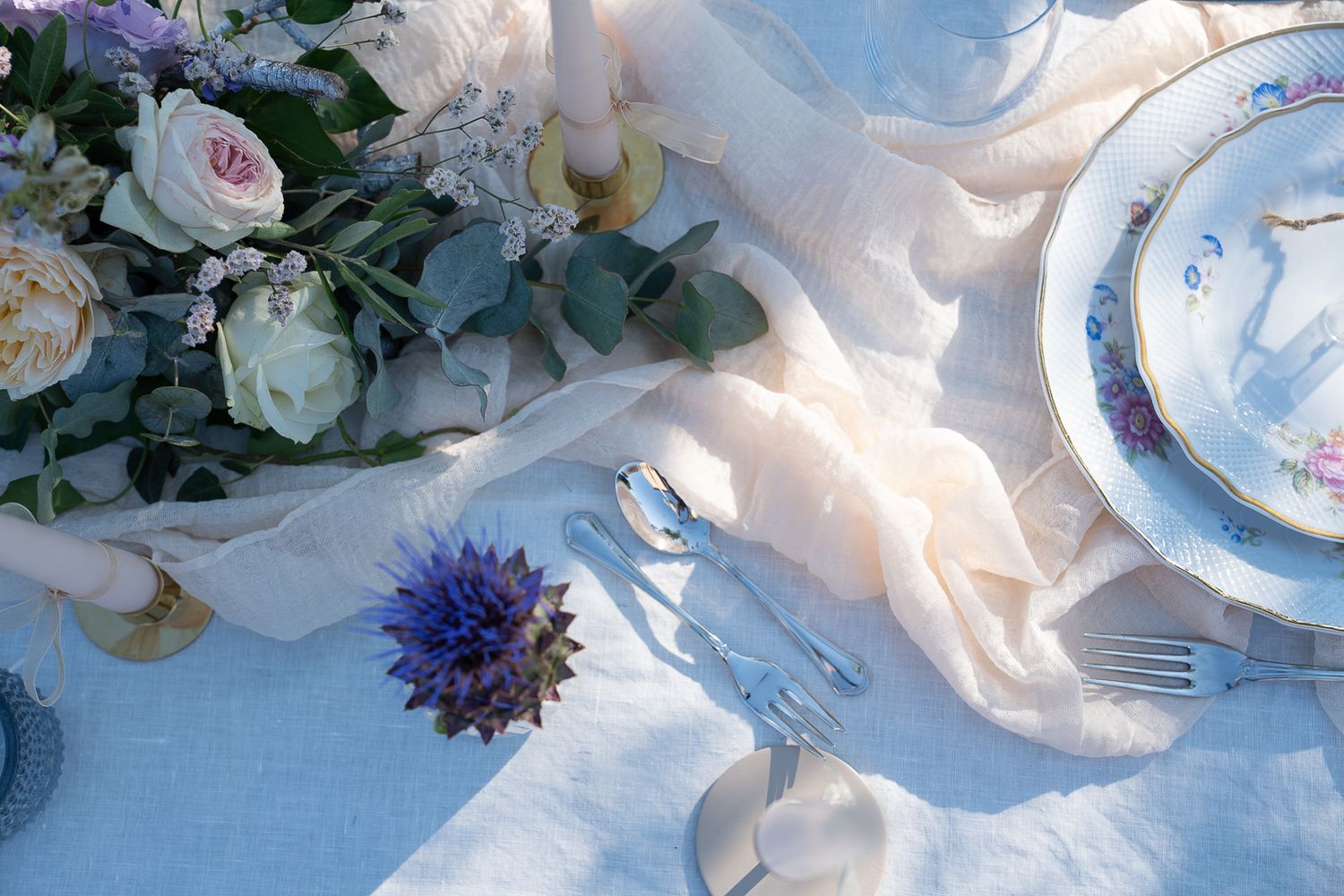 Elegant wedding table setting with floral centerpiece, fine china plates, silver cutlery, and soft linen on a blue tablecloth.