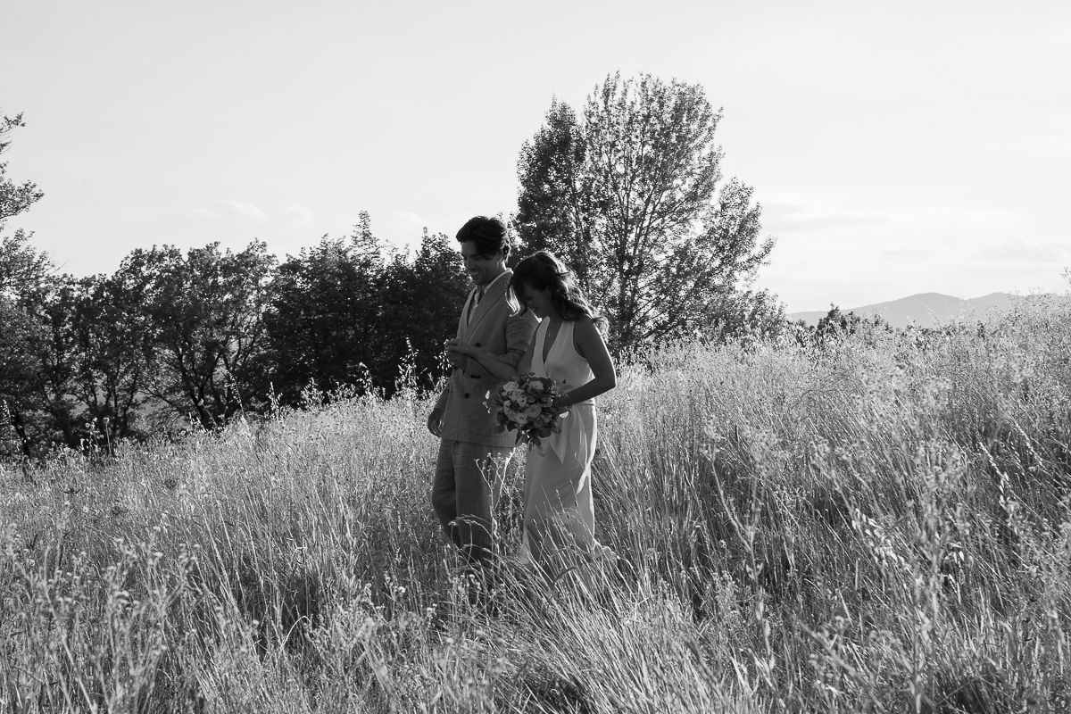 Black and white photo of a couple standing in tall grass fields with trees and mountains in the background.