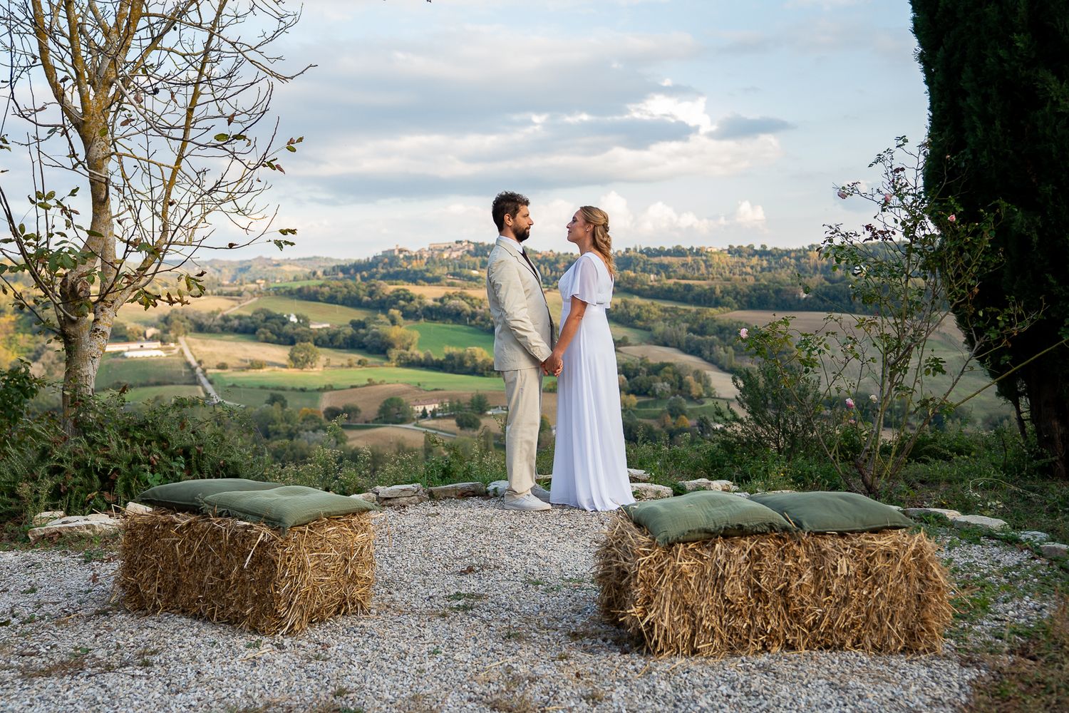 Couple in white wedding attire holding hands among hay bales with scenic Tuscan countryside in background.