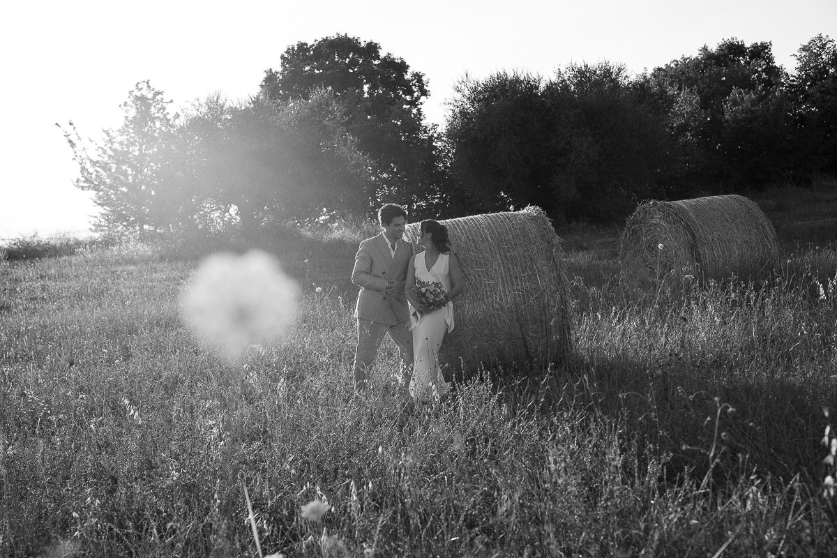 Black and white photo of a couple holding hands in a field with hay bales and trees in soft sunlight.
