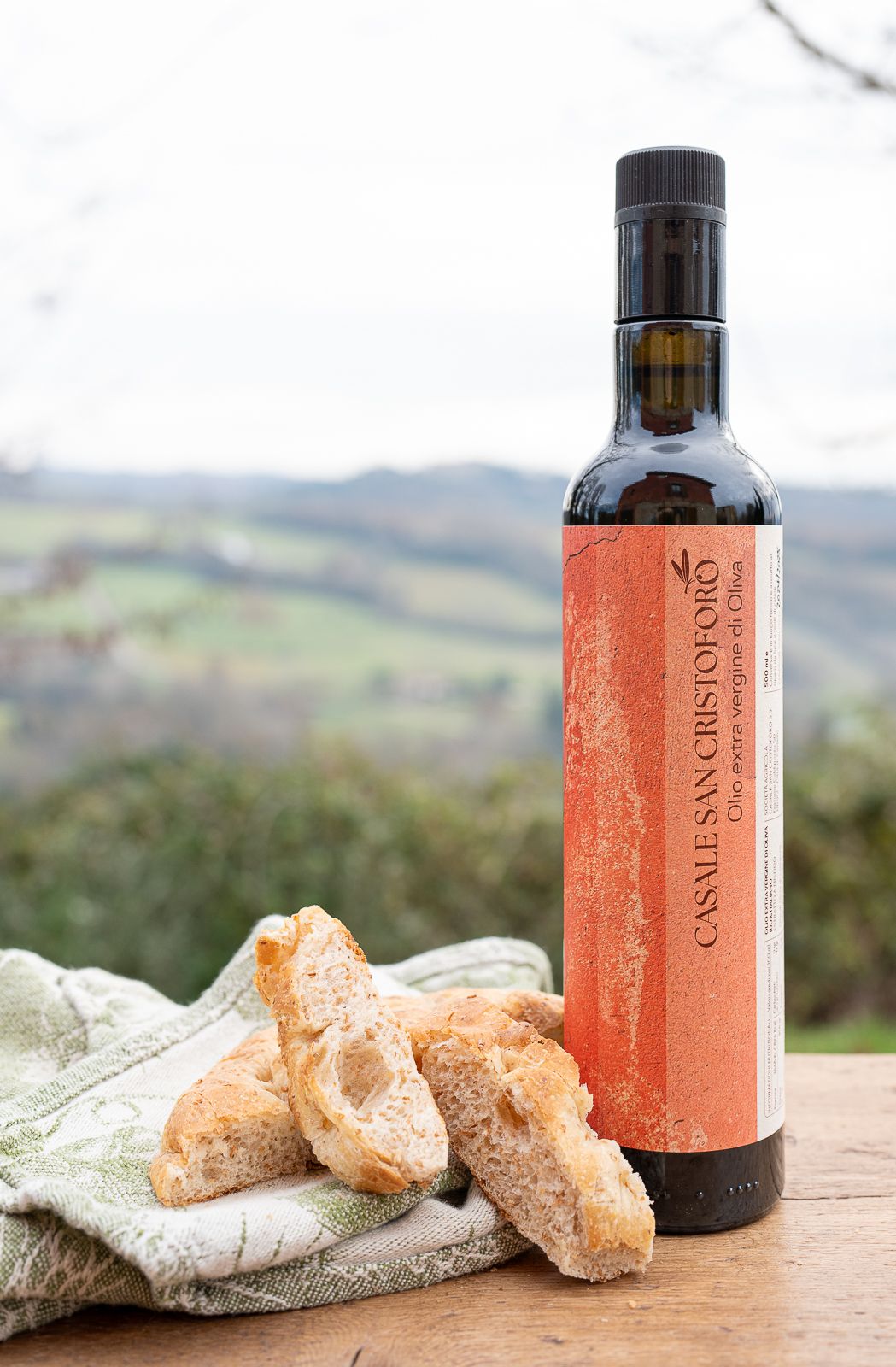 Casale San Cristoforo extra virgin olive oil bottle with rustic bread on a wooden table, Italian countryside in background.