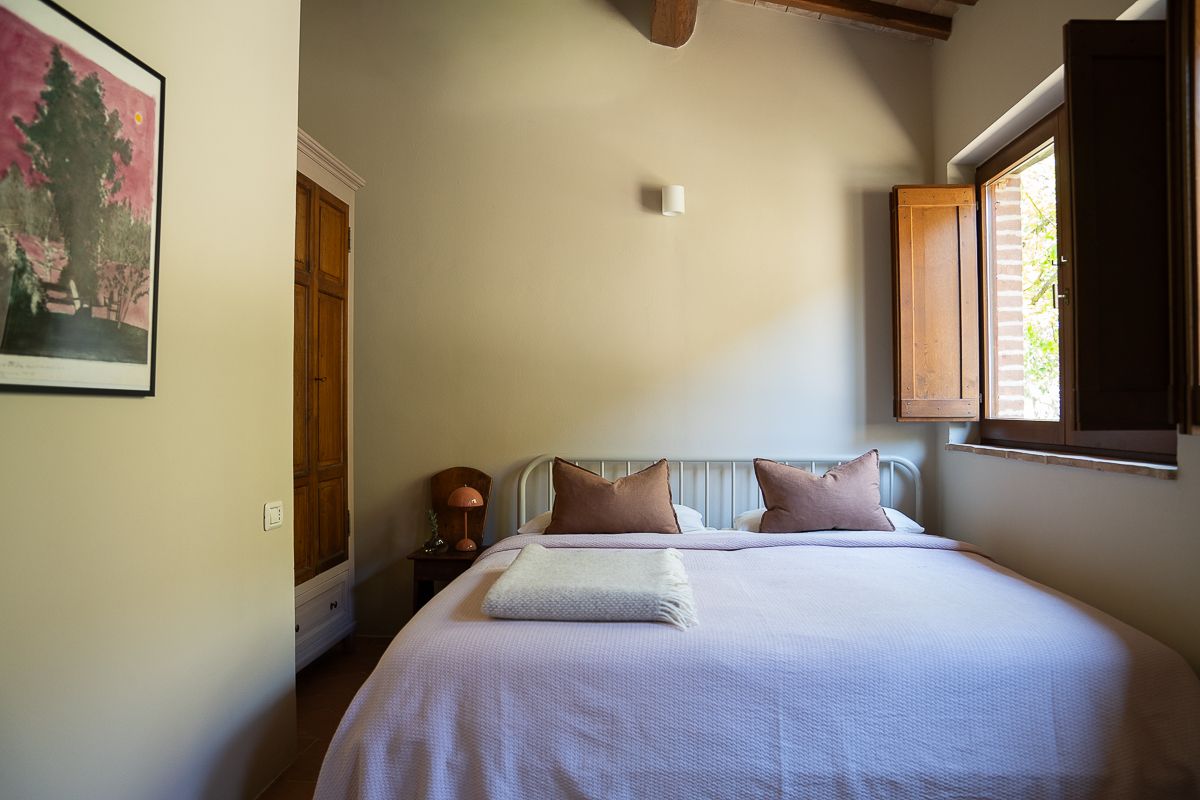 Cozy bedroom with white bedding, pink accent pillows, wooden door, wall art, and natural light from a rustic window.