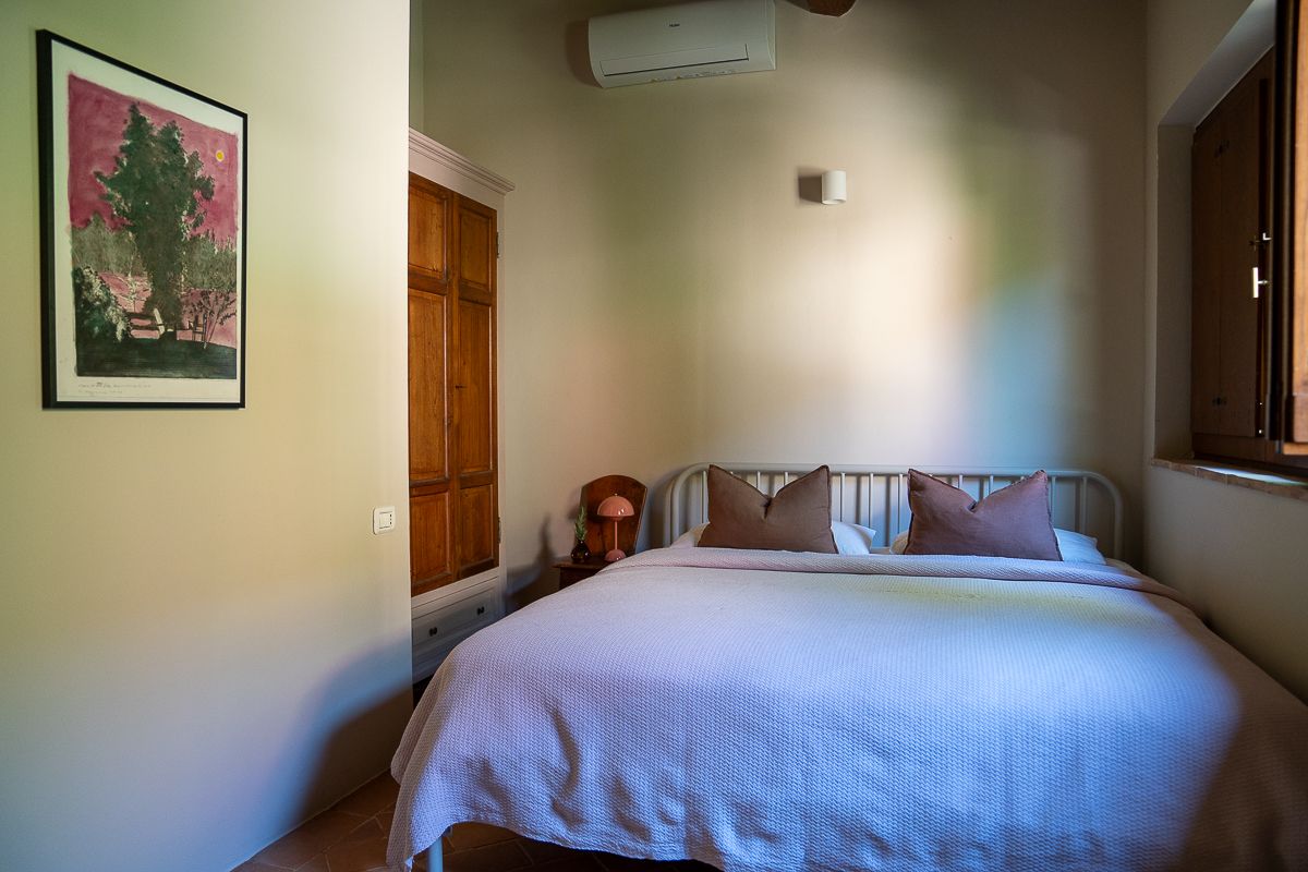 Cozy bedroom with white bedding, wooden door, wall-mounted AC unit, decorative artwork, and natural light from a side window.