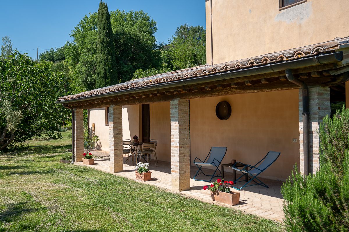 Rustic Italian farmhouse with covered terrace, lounge chairs, terracotta pots, and lush green garden surroundings.