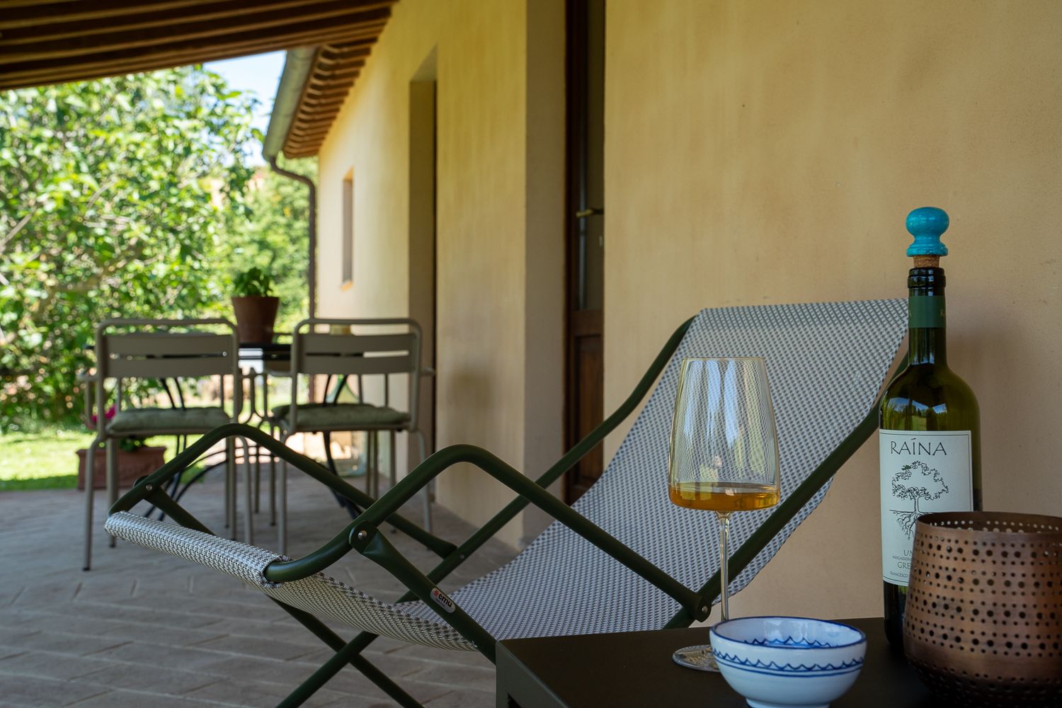 A relaxing porch scene with a lounge chair, wine bottle, and glass overlooking a garden on a sunny day.