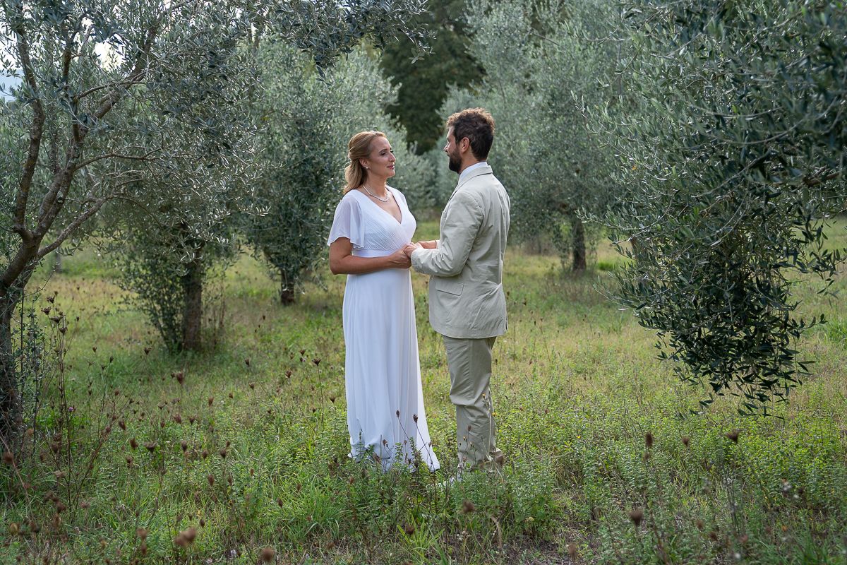 Couple walking through an olive grove on a sunny day with green grass and trees surrounding them.