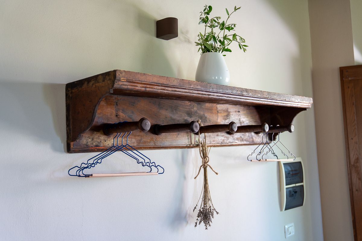 Rustic wooden wall shelf with coat hooks, hanging wire hangers, dried herbs, and a green plant in a vase.