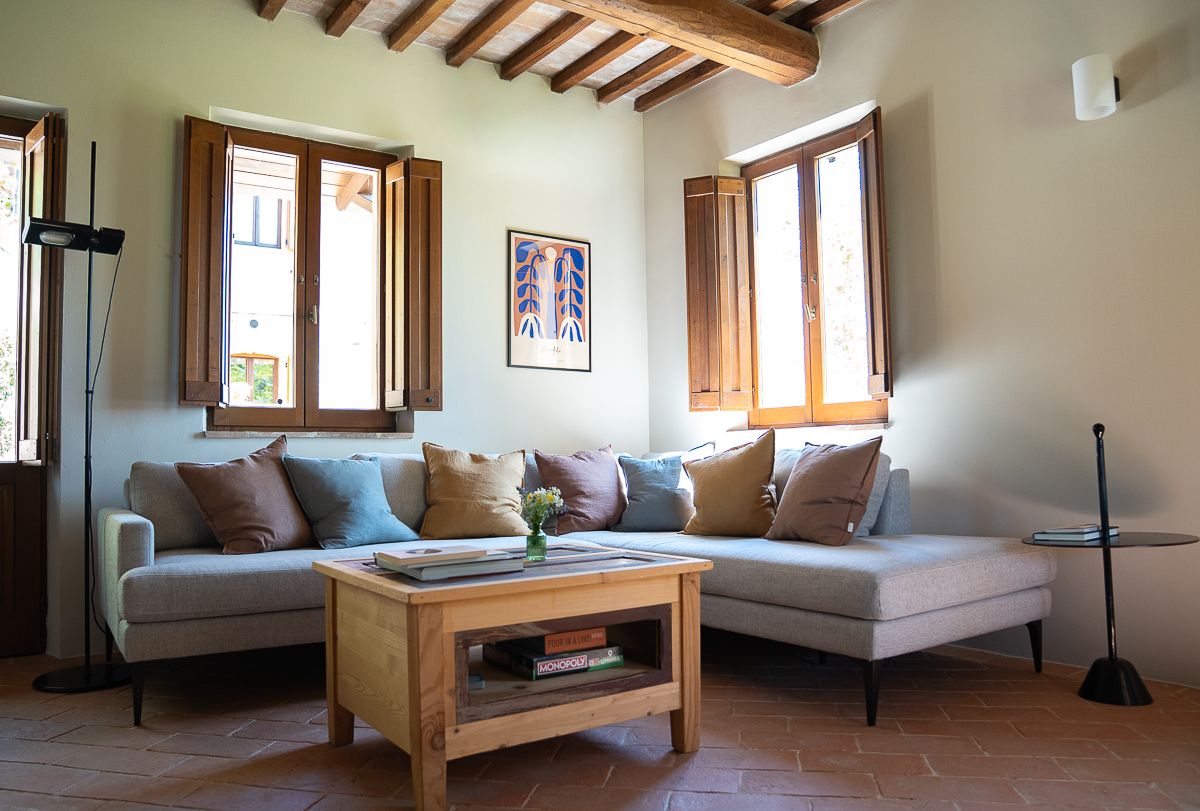 Rustic Tuscan living room with L-shaped gray sofa, wooden coffee table, terracotta floors, and beamed ceiling.