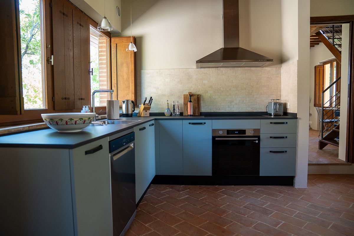 Modern kitchen with stainless steel appliances, range hood, terracotta tile floors, and natural light from wooden windows.