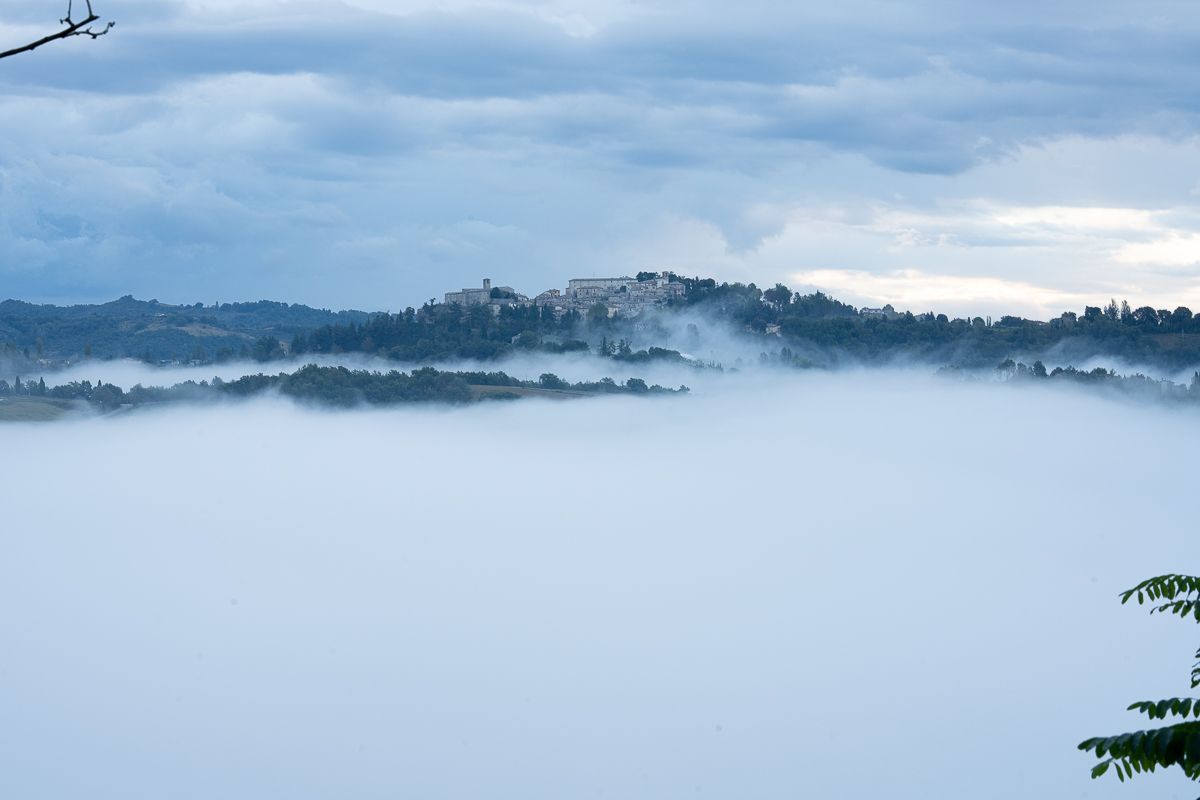 Misty valley landscape with a hilltop village emerging above a sea of fog under cloudy skies.