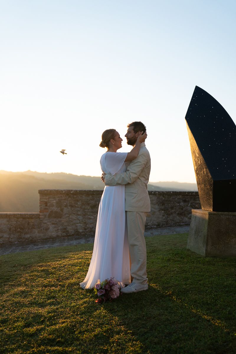 A couple in white attire embraces during sunset at a stone fortress wall as golden light illuminates the grass.