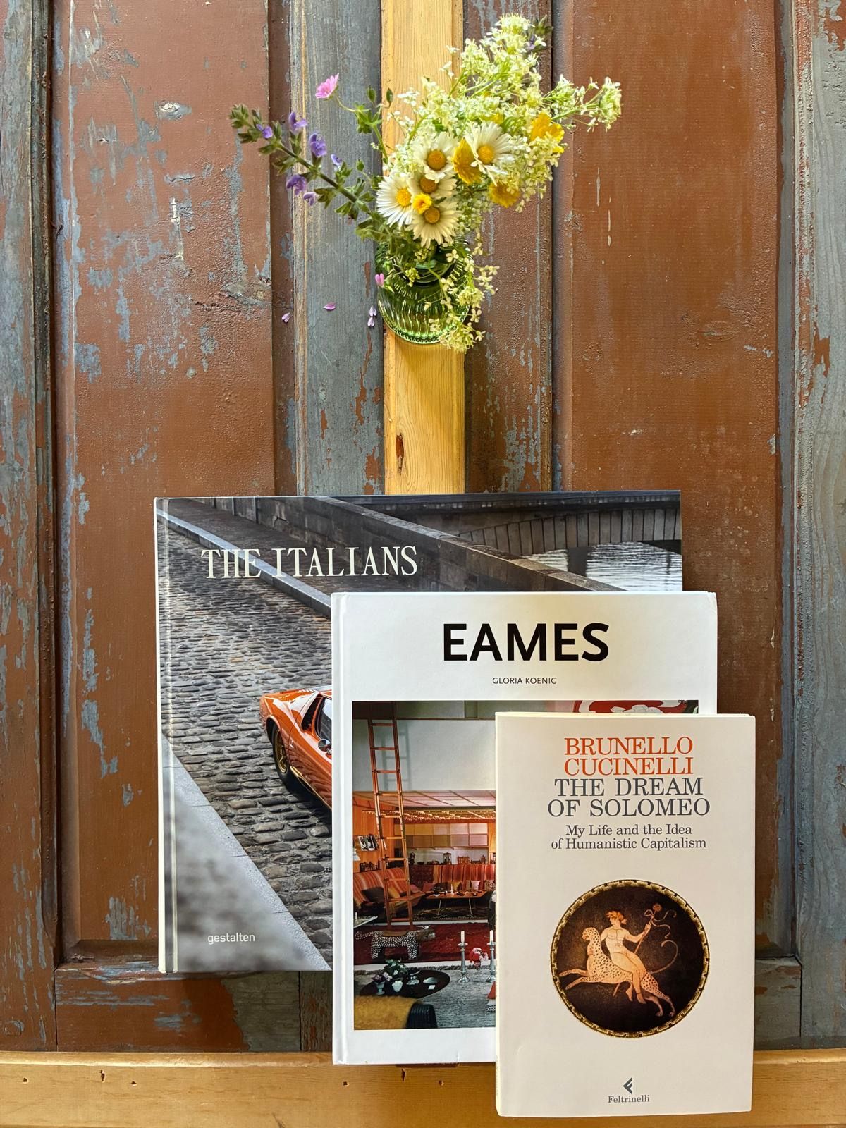 Three books including The Italians, Eames, and Dream of Solomeo displayed on wood surface with wildflower vase.