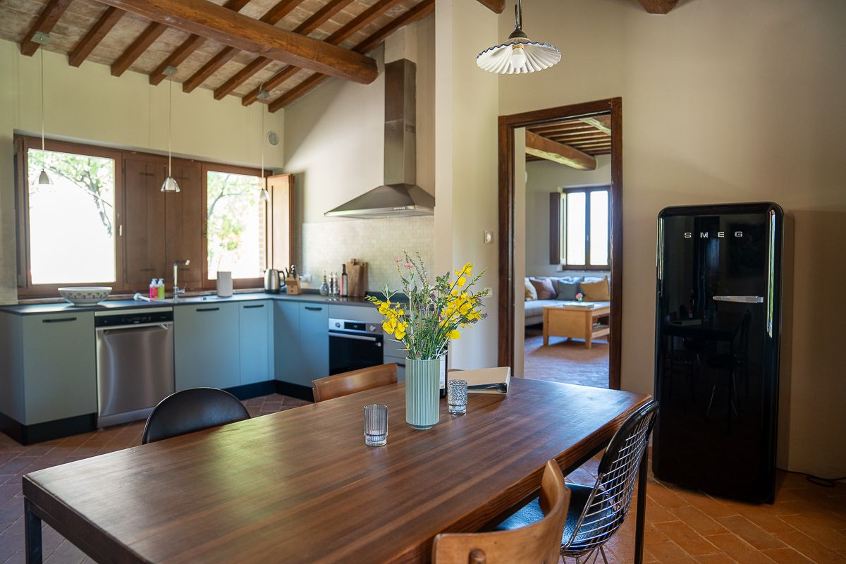 Rustic Tuscan kitchen with wooden dining table, yellow flowers, black retro fridge, and open-plan living area with terracotta floors.