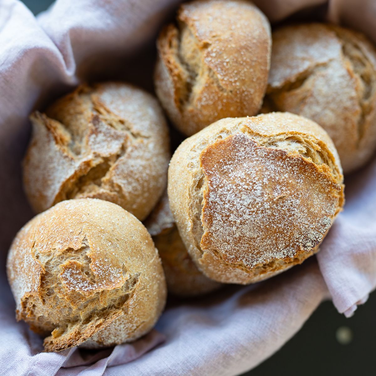 Rustic whole grain bread rolls dusted with flour, arranged in a linen-lined basket.