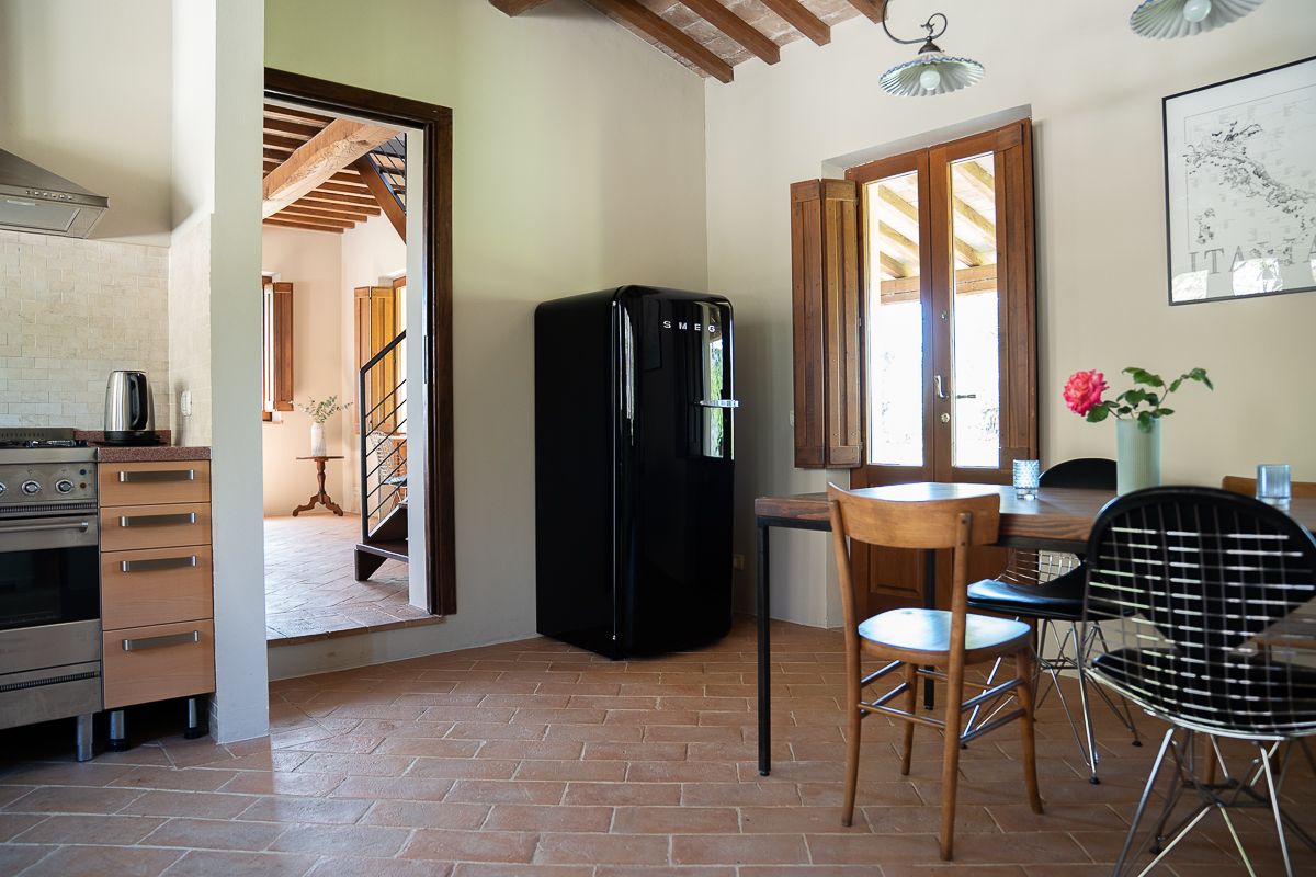 Rustic Tuscan kitchen and dining area with terracotta tile floors, black retro fridge, wooden table, and natural light from window.