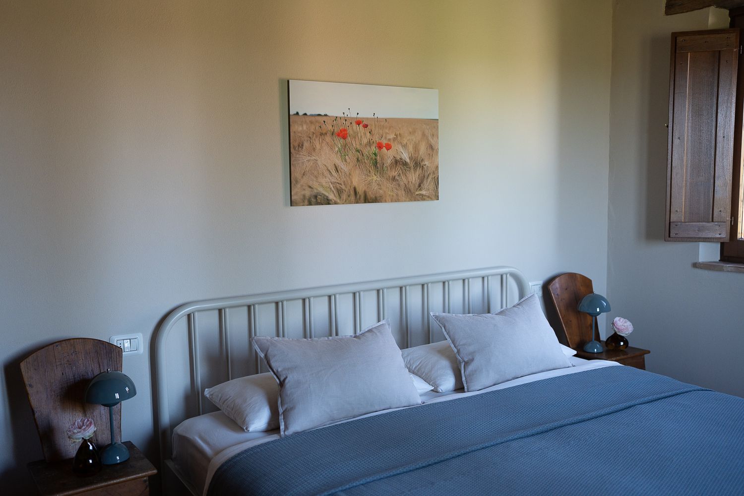 A cozy bedroom interior with metal bed frame, gray bedding, and landscape artwork on the white wall.
