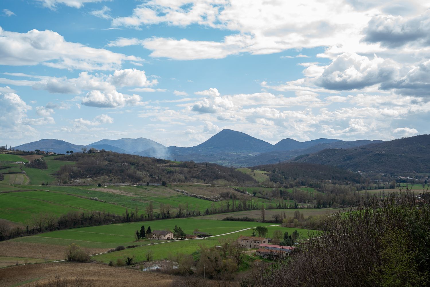 Scenic Italian countryside valley with green fields, cypress trees, farmhouse, and mountain range under a partly cloudy sky.