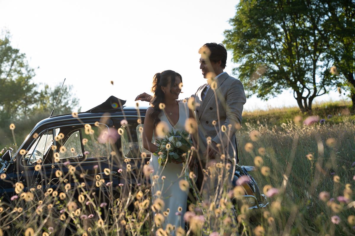 Bride and groom standing together in a wildflower field beside a vintage car at golden hour.