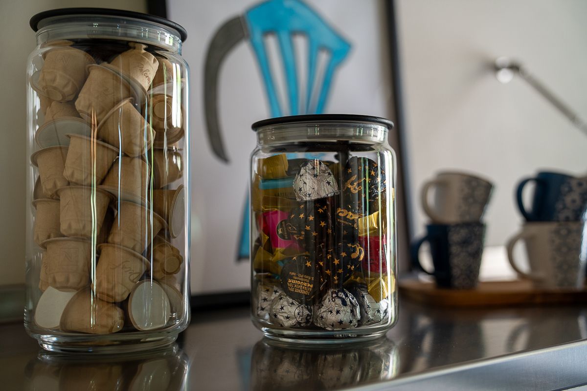 Two glass jars with lids on a countertop, one filled with corks and one with colorful items, mugs in background.